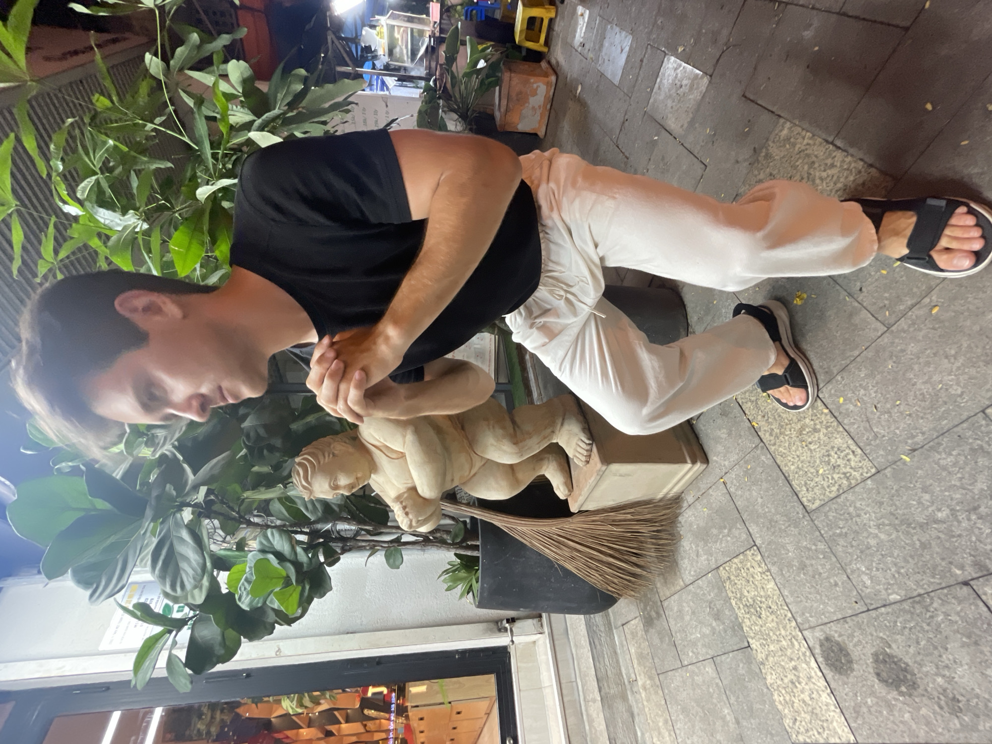 A man in a black t-shirt and white pants kneels next to a statue of a child holding a broom. The man appears to be touching the statue's hands with his hands. The statue is set within a potted plant. The location is in Thủ Đức, Vietnam as indicated by the menu in the display case. The man is wearing sandals on his feet. The scene is outdoors on a paved area.
