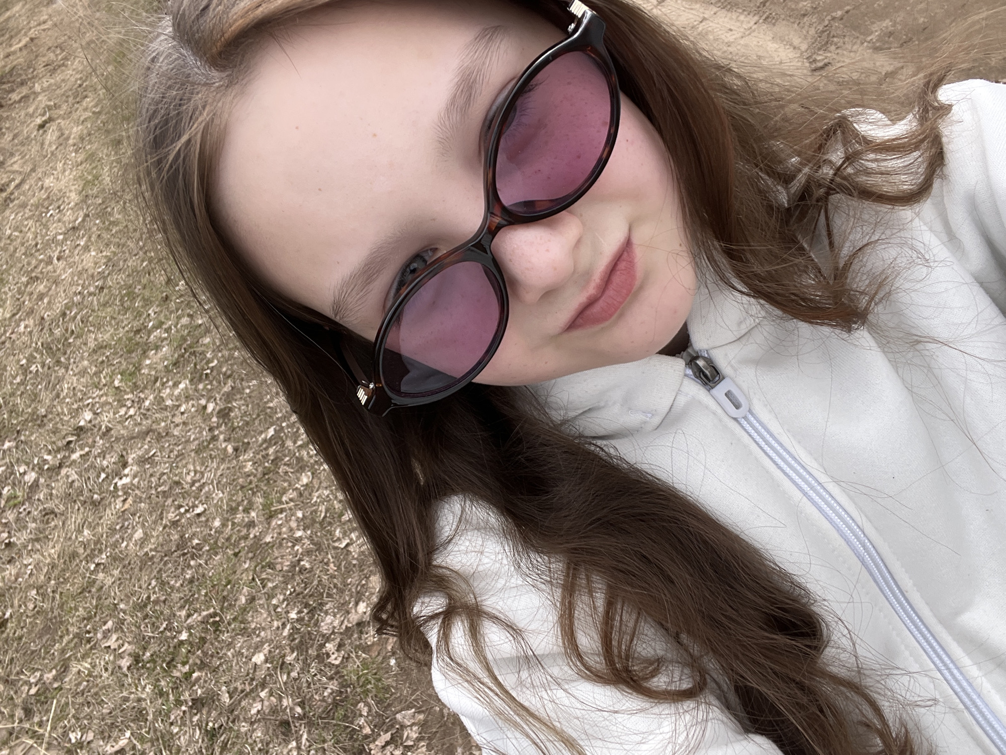 A young person wearing sunglasses and a white zip-up hoodie is posing for a selfie outdoors. The person has long brown hair. The background consists of dried grass and leaves on the ground. The location appears to be a natural area in or around the city of Гаревая, Russia.