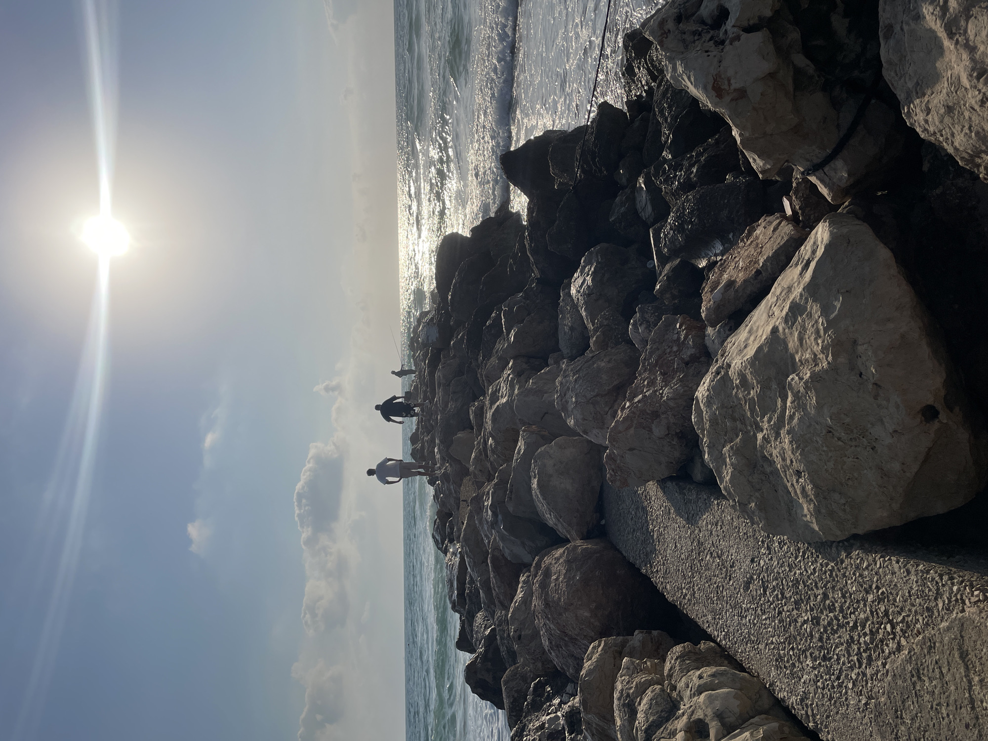 The image depicts a rocky jetty extending into the sea. The sun is bright and high in the sky, creating a glare on the water. Several individuals are visible on the jetty. Two men in casual attire are walking, one of whom is standing with his hands on his hips. Further down the jetty, another person appears to be fishing. The waves are breaking against the rocks, and the water has a choppy texture. The jetty is constructed from large, uneven stones, with a gravel pathway visible in the foreground. The scene suggests a recreational outing by the sea. This location is consistent with the coastline of Haifa, Israel, particularly areas with man-made structures like breakwaters or jetties for fishing and recreation.
