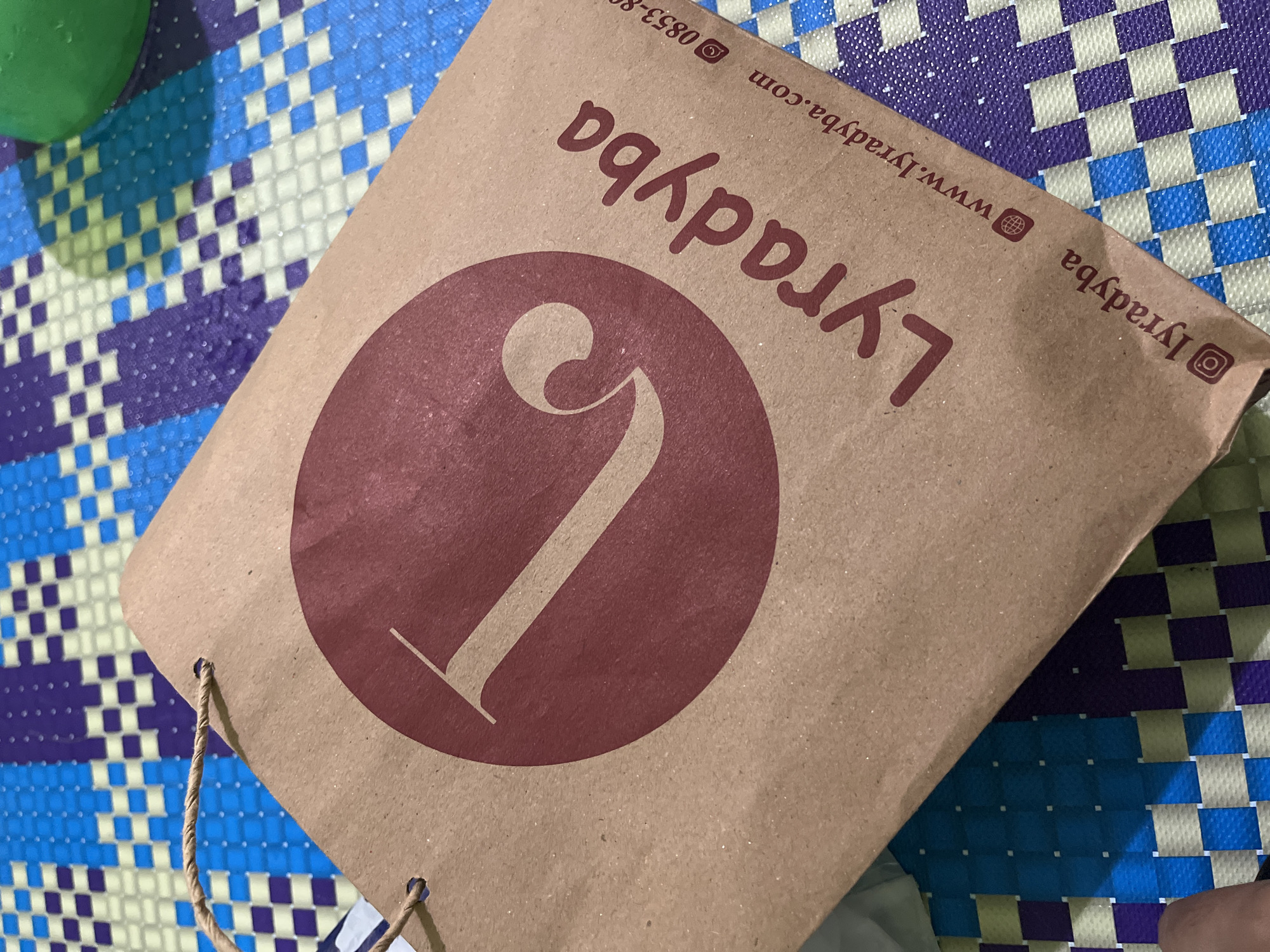 A brown paper shopping bag with a maroon circular logo containing the letter "L" and the text "Lyradyba" is placed on a mat with a blue, purple, and white checkered pattern. The bag has a twisted rope handle. To the right of the bag, a portion of a green spherical object is visible. The scene is indoors, and the lighting suggests it is daytime. There are no people clearly visible in the image. The location is the city of Babahjurong, Indonesia.