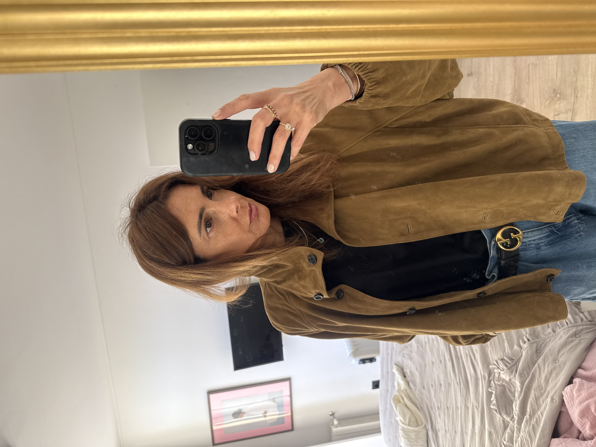 The image captures a woman taking a mirror selfie in an indoor setting, likely a bedroom or personal living space in Vila Nova de Gaia, Portugal. She is seen from the chest up, with her head slightly tilted and a neutral expression as she looks into the phone's camera. Her brown hair falls to her shoulders.
She is wearing a brown, suede-like jacket over a black top, which has faint, unidentifiable text across the chest. Below the jacket, she wears blue jeans and a black belt with a distinctive gold 'G' shaped buckle, commonly associated with Gucci. Her right hand, holding a black smartphone (likely an iPhone with multiple cameras), is adorned with several rings, including one prominent ring with a large clear stone and another with multiple bands. A thin, sparkling bracelet is visible on her left wrist.
The reflection in the large gold-framed mirror reveals a background consisting of a white wall, a mounted black rectangular screen, and a framed abstract artwork featuring pink and grey tones. Part of a bed with white and pink bedding and a white suitcase are also visible, along with a light-colored wooden floor. The overall lighting suggests it is daytime.