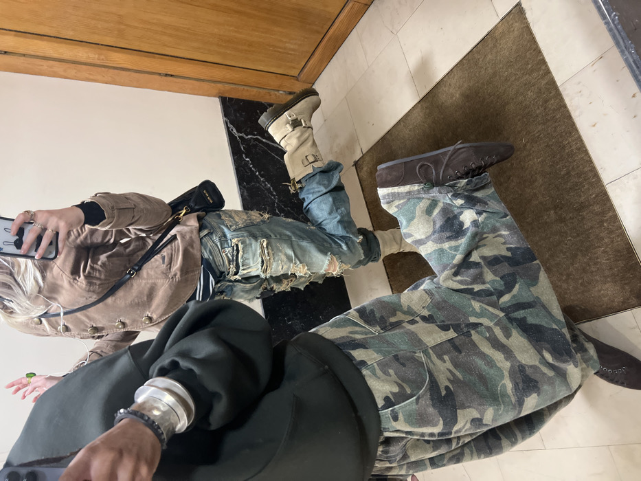 The image, rotated 90 degrees clockwise, depicts a mirror selfie or reflection of two individuals, primarily showing their attire and a portion of the indoor setting.
On the left side of the corrected image, a person with blonde hair is visible, appearing to take the photo with a black smartphone held in their left hand. The phone has a white charm or detail on its case. This person is wearing a light brown or tan jacket, possibly made of suede or a similar textured fabric, featuring multiple buttons down the front and flap pockets. Underneath the jacket, a black top is visible. They are wearing heavily distressed, light blue denim jeans with significant rips and tears on the thighs and knees. Their footwear consists of light beige or cream-colored boots with two visible buckles on the side. Accessories include multiple silver rings on the fingers of the left hand, a prominent silver cuff bracelet on the right wrist, and a darker beaded bracelet next to it. A small black shoulder bag with gold hardware is slung over their shoulder, and a faint inscription that appears to read "DIOR HOME" is visible on it.
On the right side of the corrected image, a second person is standing. They are wearing a dark green or black hooded sweatshirt, with the hood visible on their upper back. Their lower body is clad in loose-fitting or baggy camouflage print pants, featuring shades of green, brown, and beige. They are wearing dark brown lace-up shoes or boots.
The setting appears to be indoors. The floor features a pattern of black and white tiles, possibly marble, near the first person, and transitions to a dark brown rug or mat under the second person. A light-colored wall (white or cream) with a horizontal wooden trim or door frame runs across the top of the image. The lighting is bright and even, suggesting an indoor environment with good illumination.
