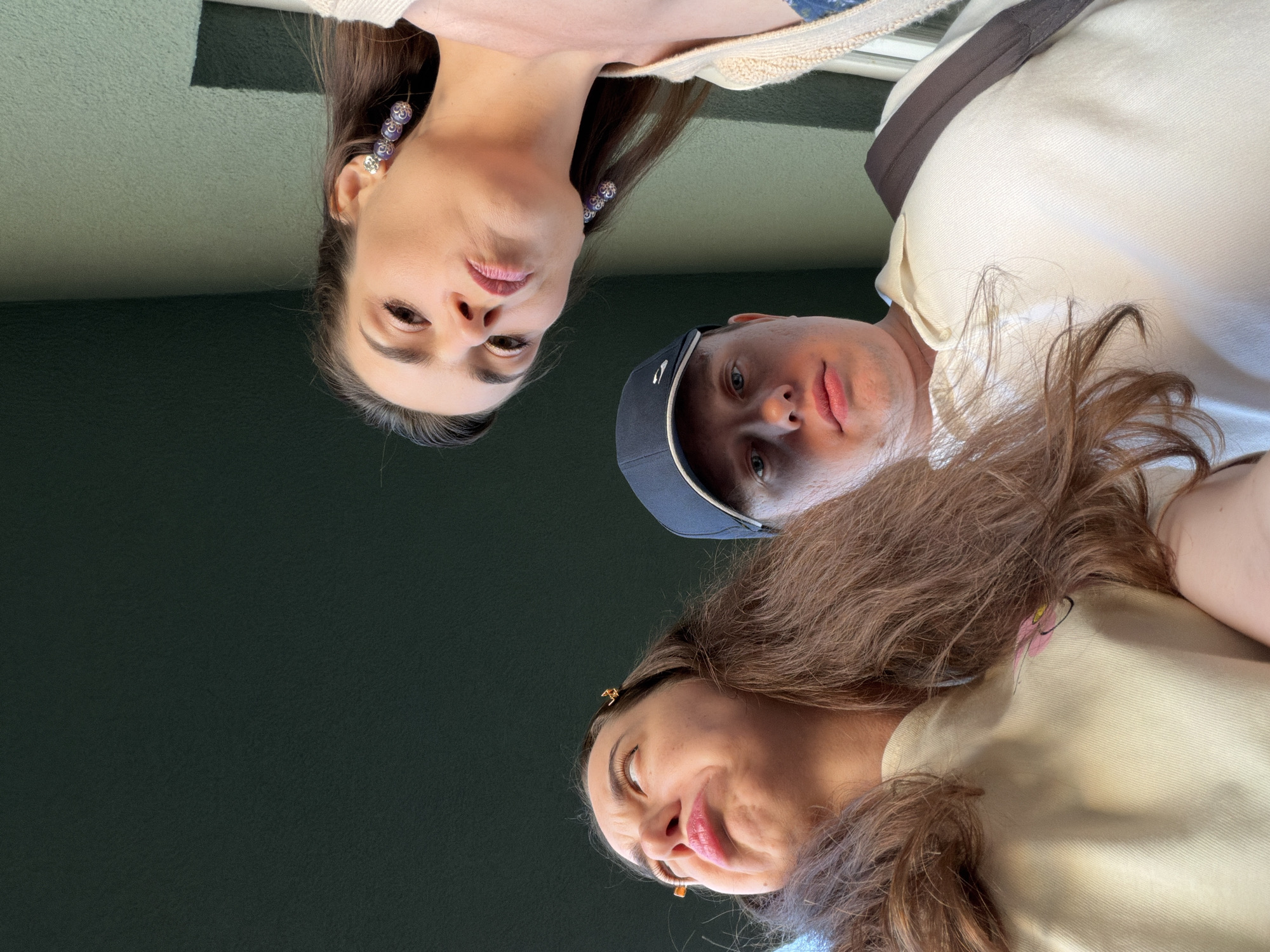 An upward-angle, low-light, medium shot captures three individuals in what appears to be an outdoor urban setting. A young woman with long, dark brown hair and a light yellow shirt is on the left, her head tilted back and her nose wrinkled in an exaggerated expression. She has small golden hair clips in her hair. In the center, a young man wearing a dark blue baseball cap and a light-colored polo shirt looks directly at the camera with a neutral expression. To the right, another young woman with dark brown hair and distinctive purple beaded earrings leans into the frame, her gaze directed downward with a stern expression. She is wearing a light-colored cardigan over a blue garment. The background consists of textured green walls and dark overhangs, typical of building facades. The lighting suggests it is daytime, possibly late afternoon, with shadows cast by the overhead structures. No specific landmarks indicating Kyiv, Ukraine are visible.
