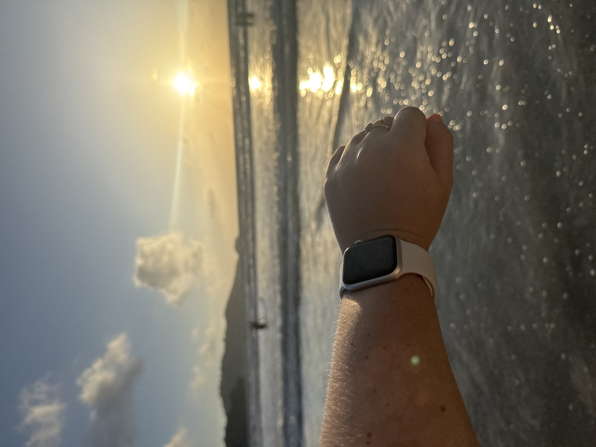 An outdoor, eye-level, close-up shot captures a person's arm and hand wearing a silver Apple Watch with a white band. The arm is positioned in the foreground, with the hand slightly clenched and a ring on the ring finger. The background features a beach scene at sunset. The sun is low on the horizon, casting a warm glow and reflecting off the ocean waves. The water is a deep blue with white foam from the surf. In the distance, a person can be seen wading in the water, and a silhouetted island or landmass is visible against the sky. The sky is a gradient of blue and orange, with scattered clouds. The location is implied to be Patong, Thailand, given the context provided.