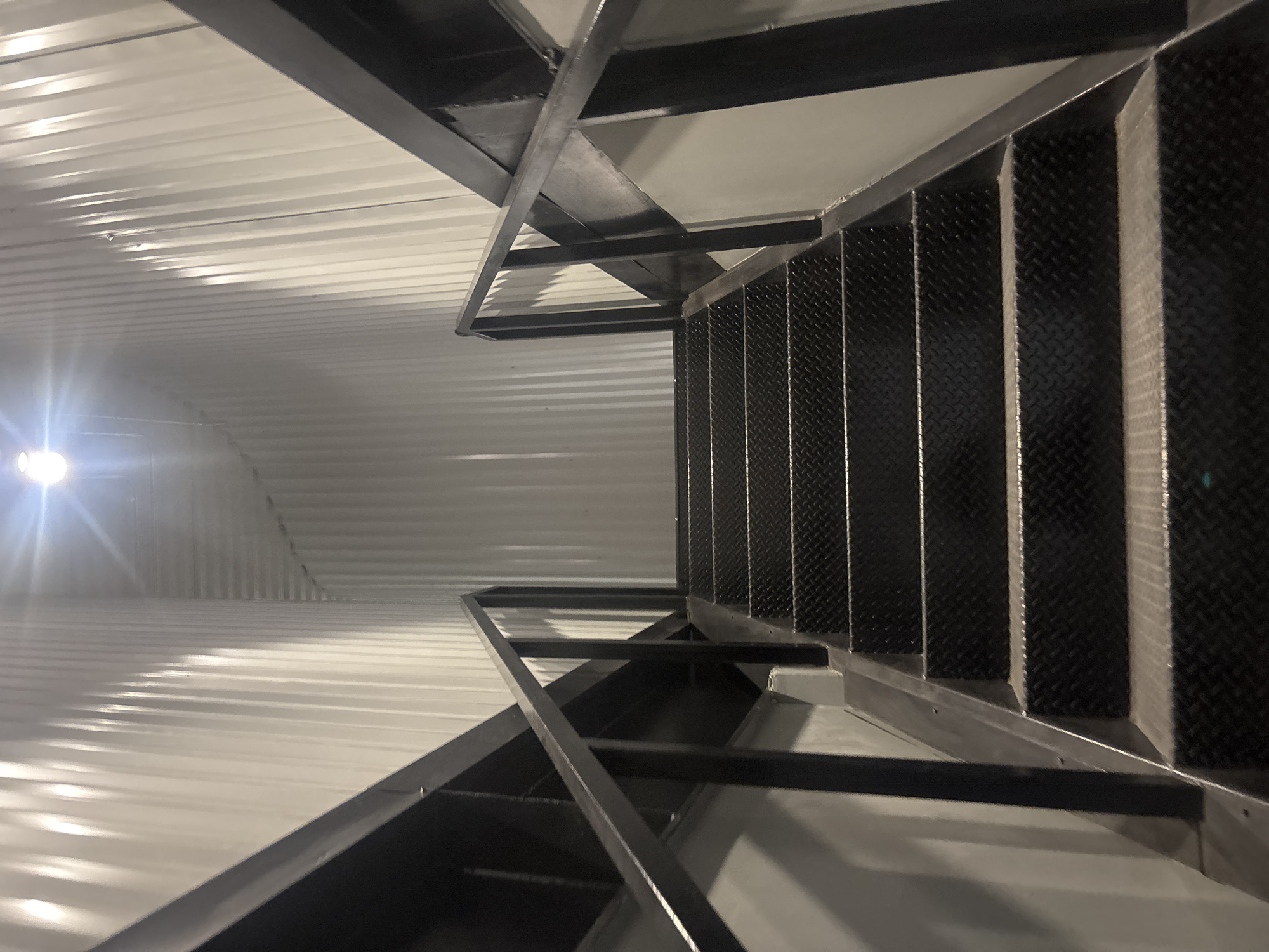 This image captures an upward view of a stark, industrial staircase within an indoor setting, likely in Buenavista, Mexico. The primary subjects are a dark metal staircase and a corrugated metal wall or ceiling.
The stairs are constructed from dark, possibly black, metal with textured diamond plate treads, indicating an industrial or utility design. They ascend steeply, supported by a sturdy framework of dark metal beams and angled braces that also form the surrounding structure. The handrails are integrated into this metal framework.
To the left, a corrugated metal surface, painted white or light grey, extends into the background. This surface is brightly illuminated by a very strong, unshielded light source positioned in the lower-left. The light creates prominent reflections and shadows that accentuate the ribbed pattern of the corrugated material, and a noticeable lens flare is present. The contrast between the bright, reflective wall/ceiling and the darker, textured stairs is a notable visual element.
No people or activities are visible, lending a sense of quiet functionality to the scene. The intense artificial lighting suggests an interior space, likely without natural light, such as a warehouse, factory, or commercial building. The overall aesthetic is utilitarian and robust. There is no discernible text in the image.