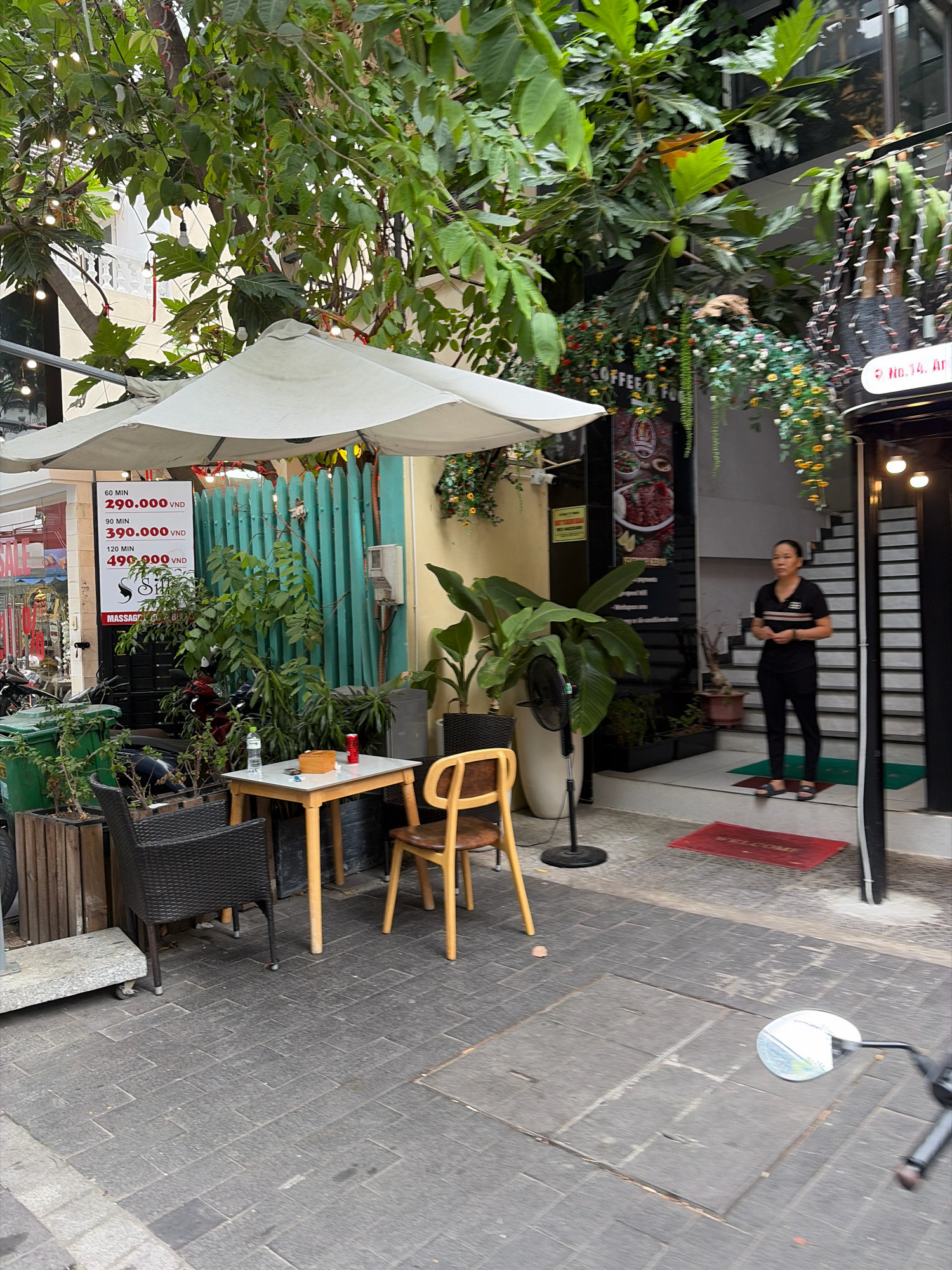 The image shows an outdoor seating area in front of a building. A white umbrella shades a small wooden table with two chairs, one wicker and one wooden. On the table, there are a bottle of water, a can of soda, and a small wooden box. A price list for massage services is displayed on a teal-colored vertical grate behind the table. A woman in black clothing stands to the right of the frame, near the entrance to the building, which has a set of stairs leading up. Lush greenery, including large-leafed plants and hanging vines with flowers, adorns the facade of the building and the surrounding area. The ground is paved with gray rectangular tiles. The scene appears to be in a commercial district, likely in Đà Nẵng, Vietnam, given the currency displayed on the price list (VND).