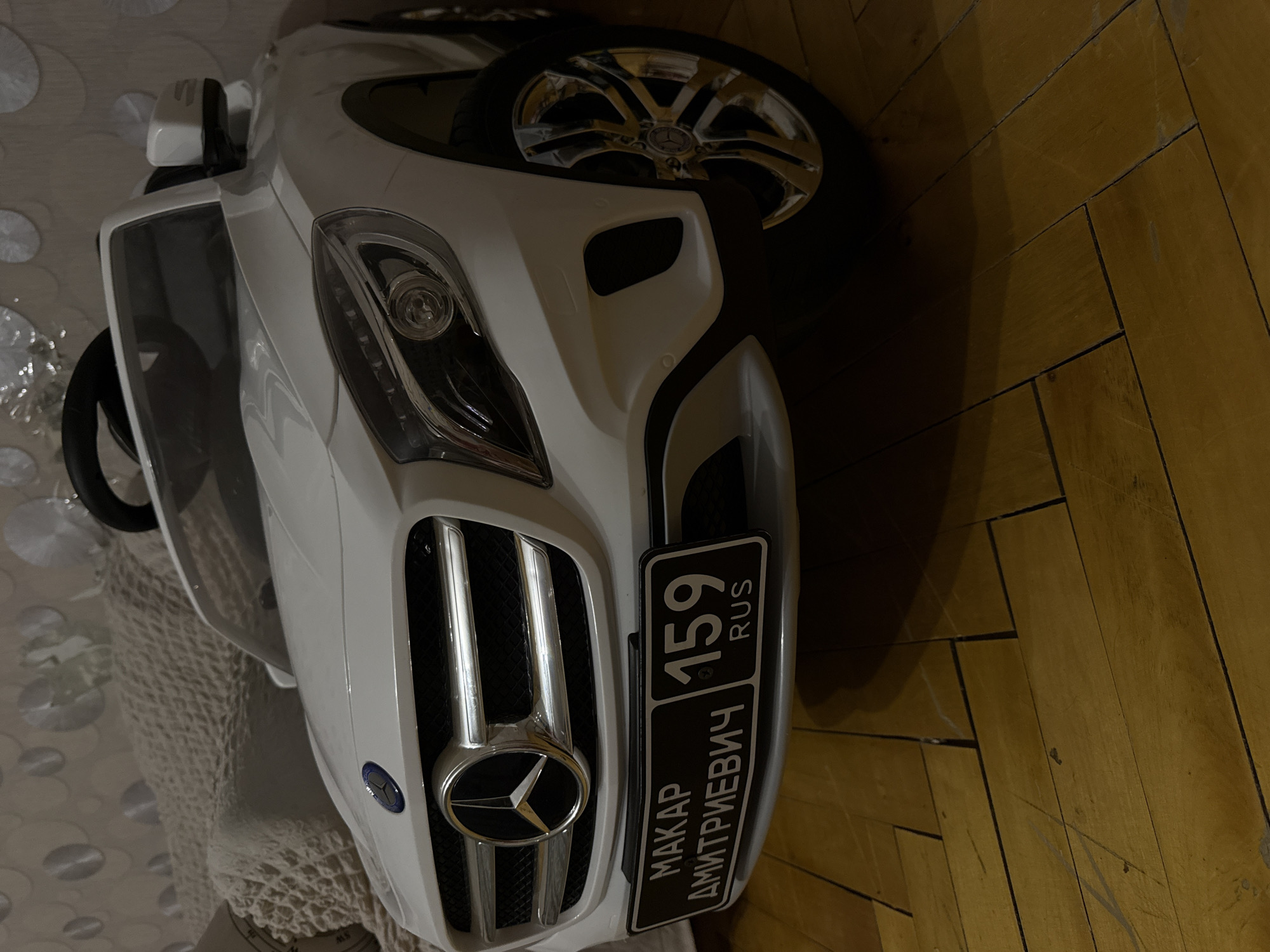 The image depicts the front of a white toy Mercedes-Benz car, positioned indoors on what appears to be a wooden floor near a patterned rug or cover. A license plate reading "MAKAR DMITRIEVICH 159 RUS" is prominently displayed. The car is partially visible, showing the front grill, headlight, and a portion of the wheel.