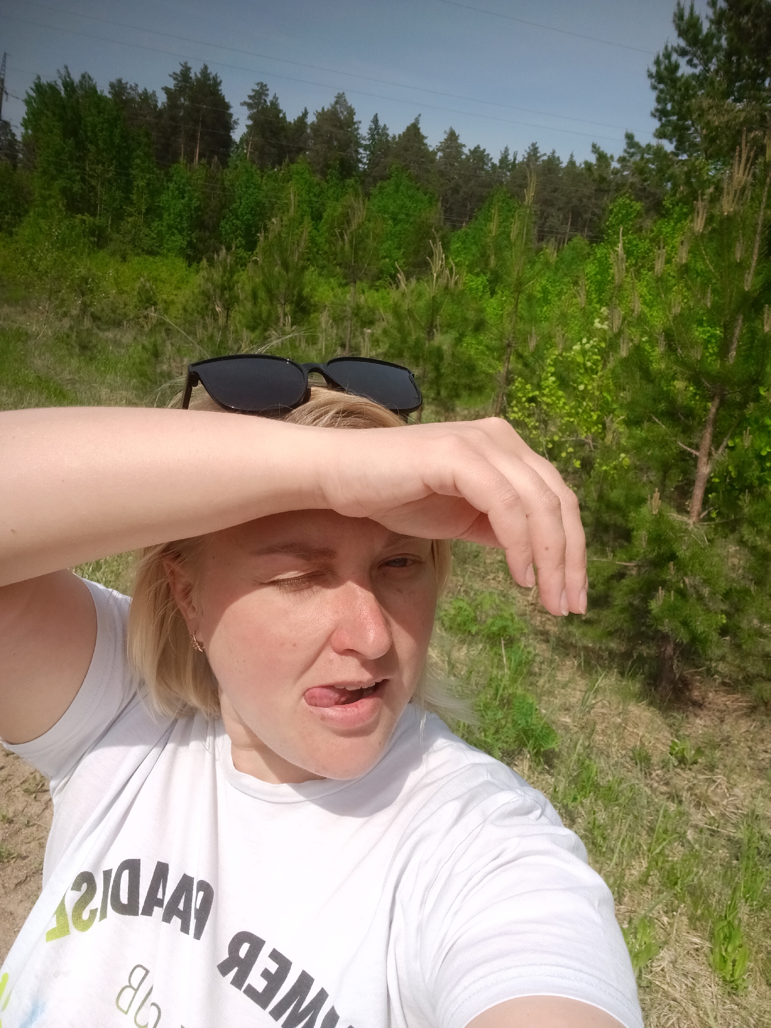 A woman with blonde hair and sunglasses perched on her head stands outdoors in a grassy area dotted with young pine trees. She is wearing a white t-shirt. Her left hand is raised to her forehead, shading her eyes, and she is sticking her tongue out. Behind her, there's a dense forest of taller trees. The location is assumed to be in or near Chaikovsky, Russia, given the prompt's instructions.