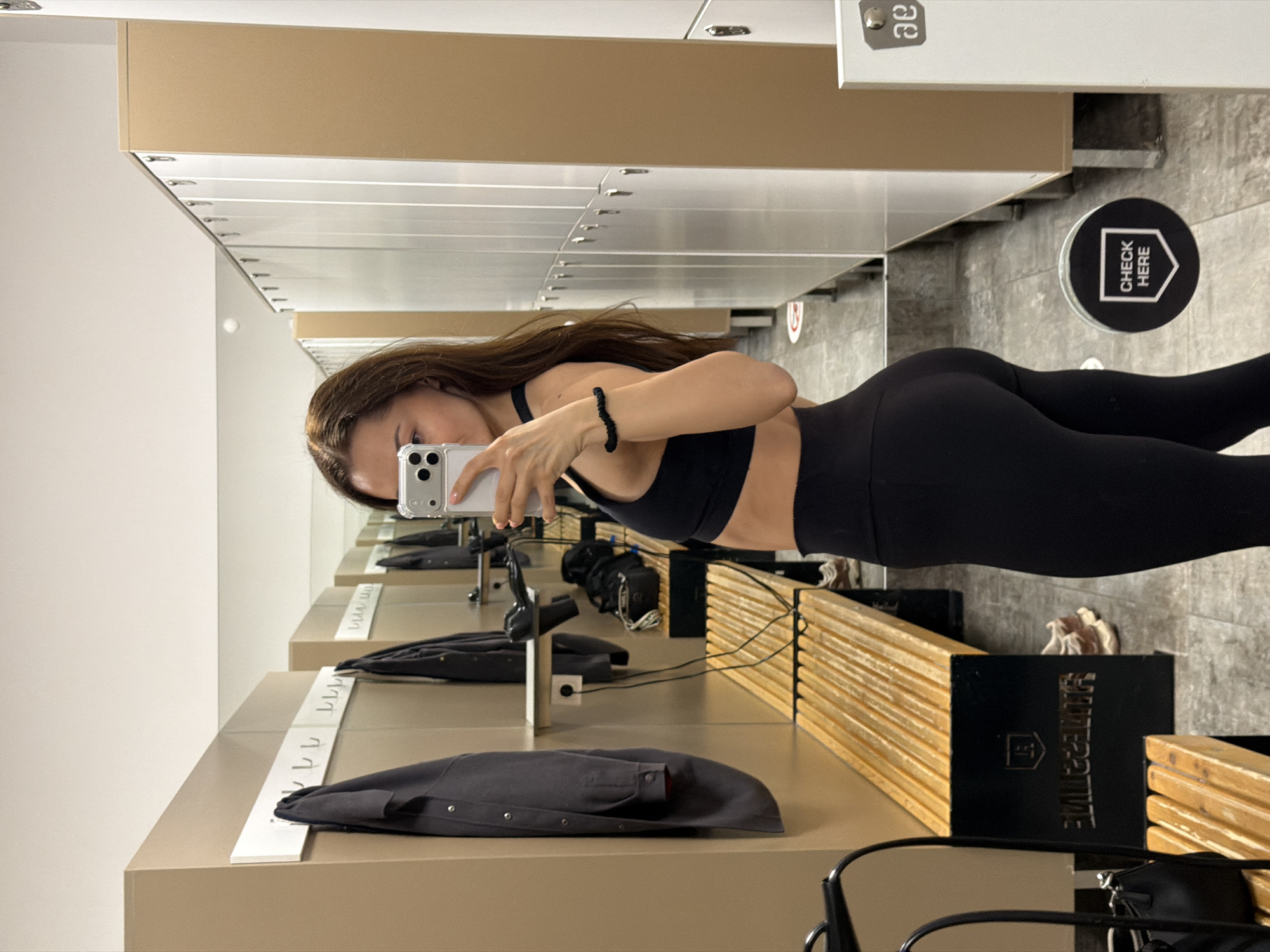 A woman is taking a selfie in a mirror, likely in the changing area of a gym. She is wearing a black sports bra and leggings. Lockers are visible behind her and to the left. There is a counter with coat hooks and clothing on it in the foreground. A "CHECK HERE" sign is visible on the floor in the background. The fitness gym is located in Varna, Bulgaria.