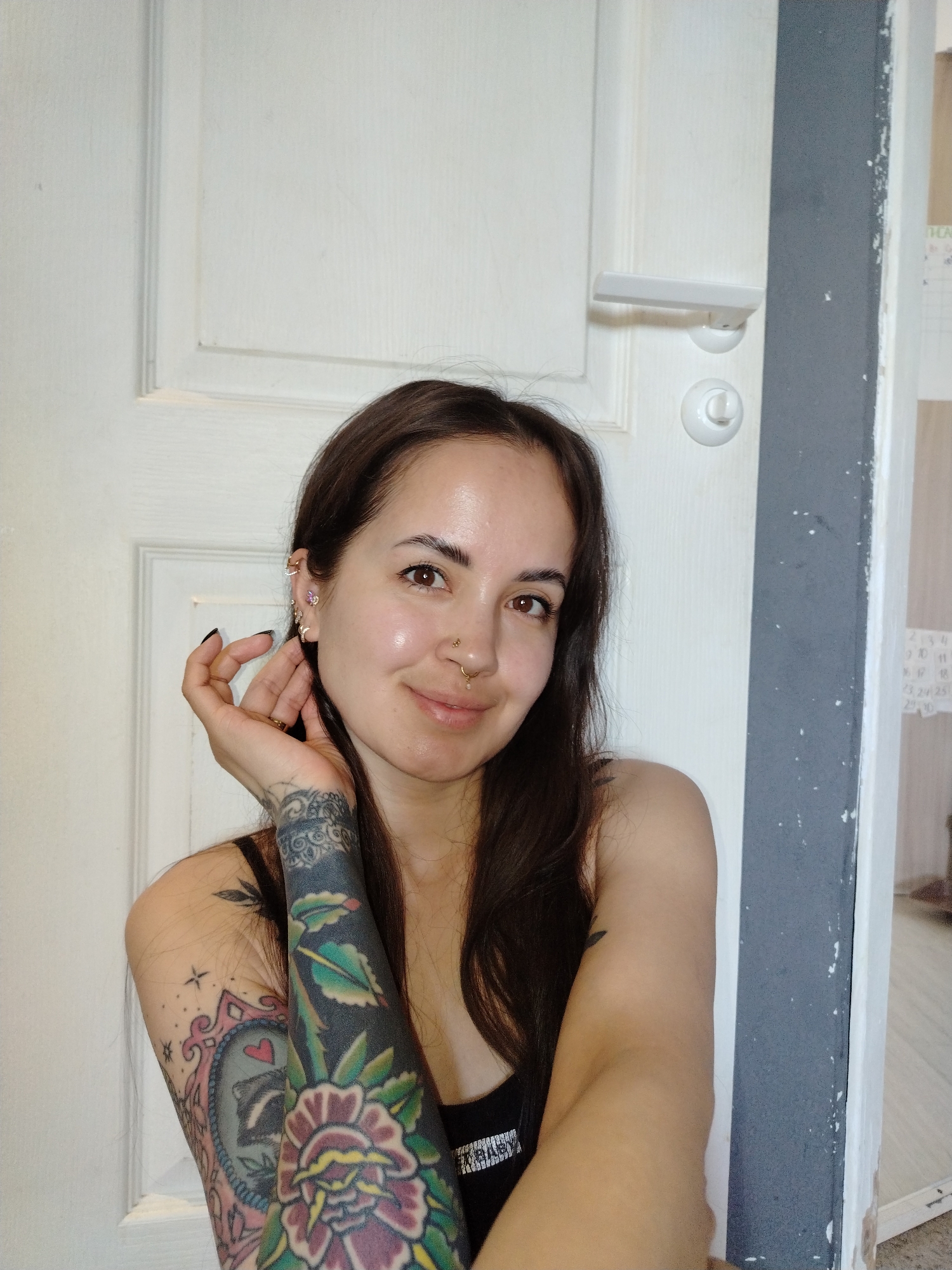 A woman with tattoos on her left arm poses in front of a white door with a white handle. The door is bordered by a gray frame on the right. A partial view of another room is visible to the right of the frame. The woman has brown hair and several piercings in her left ear and a nose ring. She is wearing a black tank top. Her left hand is raised near her face. The location in the city of Noviy, Russia, is not discernible from the image.