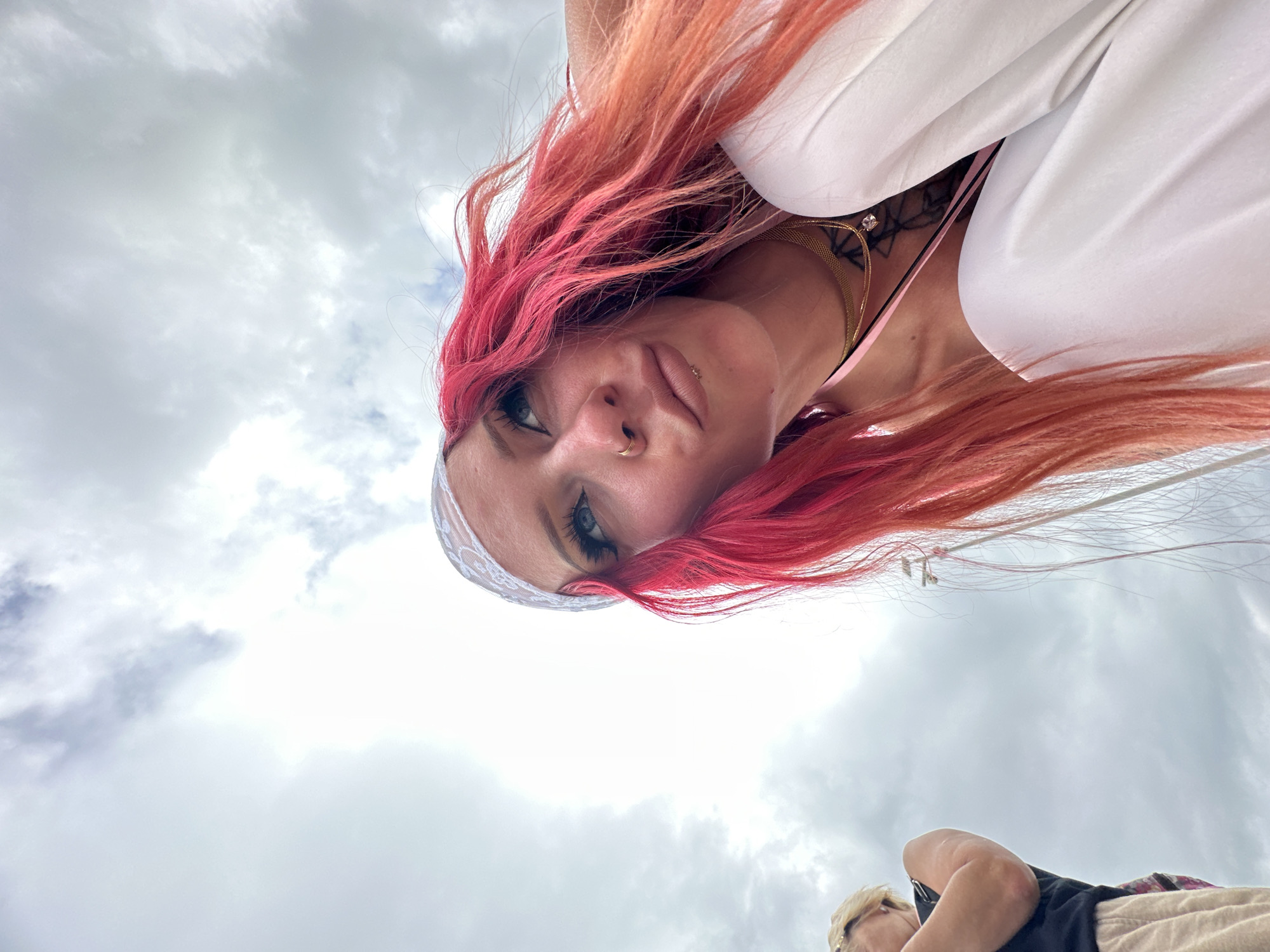A low-angle shot shows a woman with bright pink and orange hair looking down at the camera. She has fair skin and is wearing a white lace headband, a white top, and a gold necklace. A small tattoo is visible on her chest. Her expression is slightly amused. In the background is a cloudy sky. In the far bottom, there is a partial view of another person lying down. The specific location in Nusa Penida, Indonesia, is not discernible from the image alone.