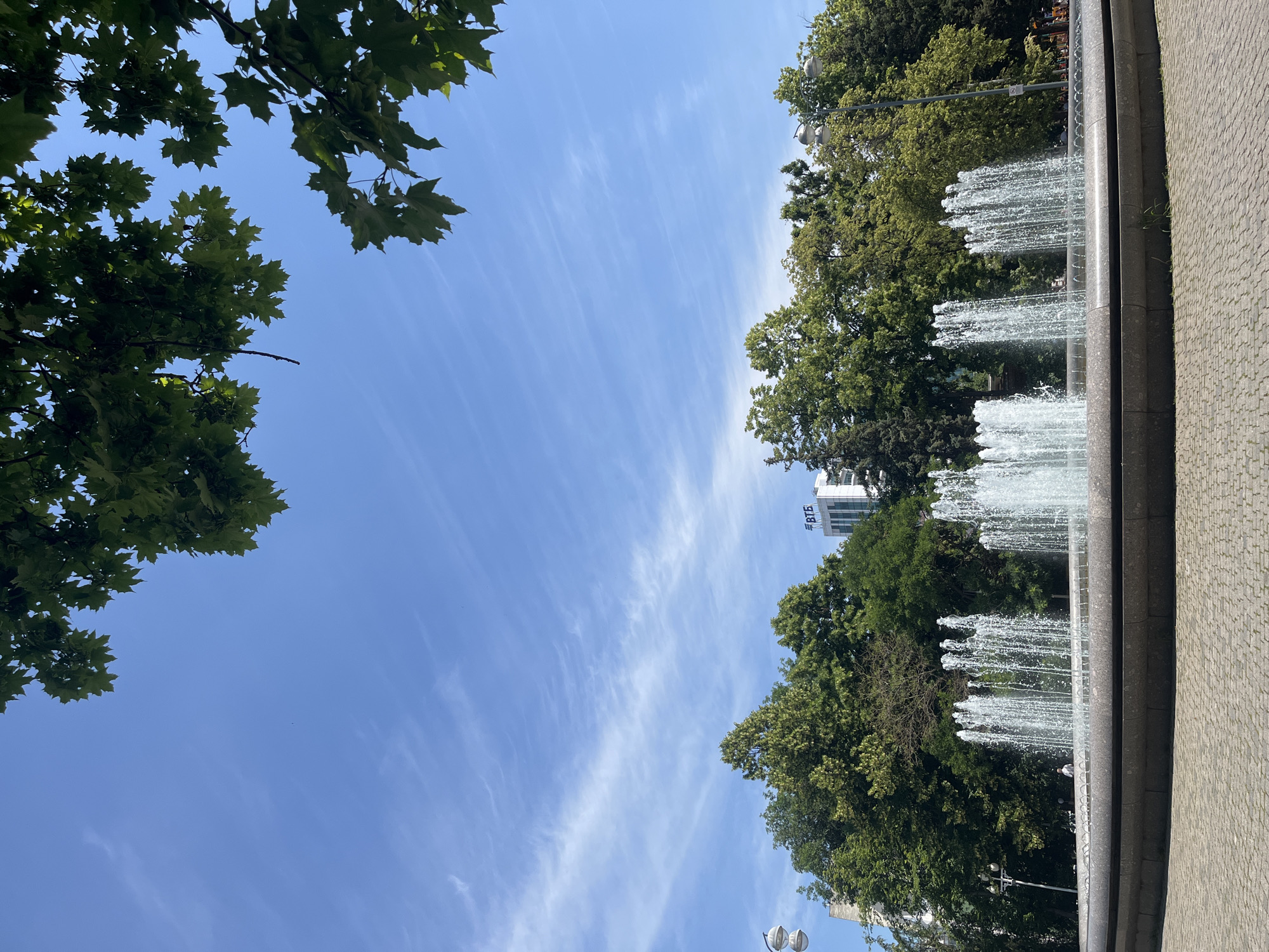 This image depicts a fountain in a city park on a clear day. The fountain is circular with multiple jets of water spraying upwards. Behind the fountain, a line of lush green trees forms a dense backdrop. In the distance, a tall white building with a blue logo that appears to be "ВТБ" is visible above the trees. The sky is a vibrant blue with thin, wispy white clouds. In the foreground, the ground is paved with cobblestones. Green leaves from overhead trees frame the top of the image. No people are clearly discernible in the foreground, though there might be faint figures in the distance within the park. The overall scene is one of a tranquil urban park setting.