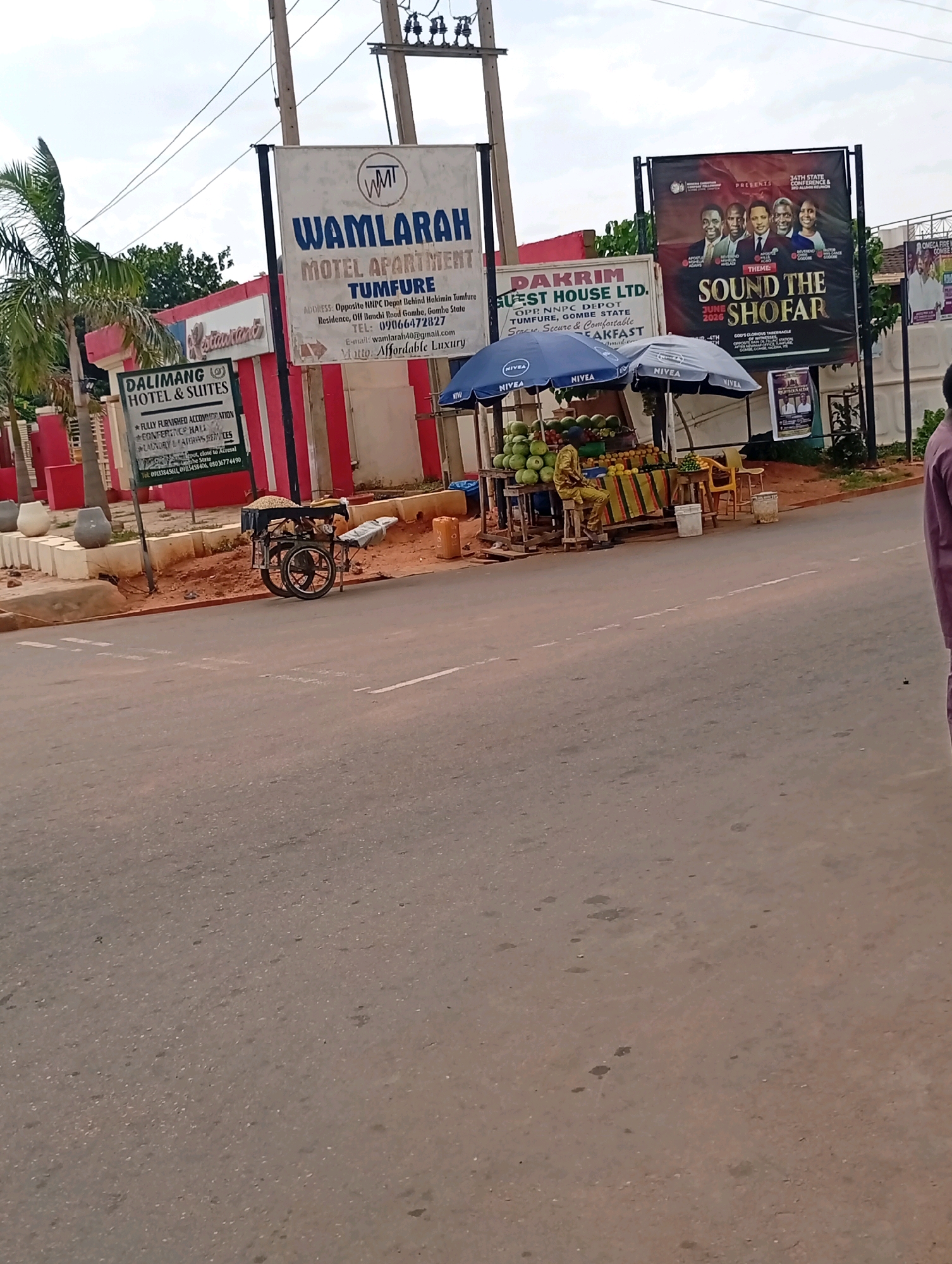 The image depicts a street scene in Tumfure, Gombe State, Nigeria, with a focus on commercial signage and roadside vendors. In the foreground, a paved road with white lane markings occupies the lower half of the frame. To the left, a sign for "DALIMANG HOTEL & SUITES" is visible, listing amenities like fully furnished accommodation and conference halls. Adjacent to this, a large banner for "WAMLARAH MOTEL APARTMENT" is prominently displayed, with an address indicating its location opposite the NNPC Depot in Tumfure.
Further to the right, another banner announces "DAKRIM GUEST HOUSE LTD." and also lists its address in Tumfure. Below these signs, a roadside stall is set up with an array of fruits, including watermelons and oranges, under blue and grey NIVEA branded umbrellas. A person dressed in a yellow outfit is seated at the stall, appearing to be a vendor. A two-wheeled cart with what looks like grains or seeds is parked nearby. In the background, more banners and buildings line the street, suggesting a commercial area. The sky is overcast.