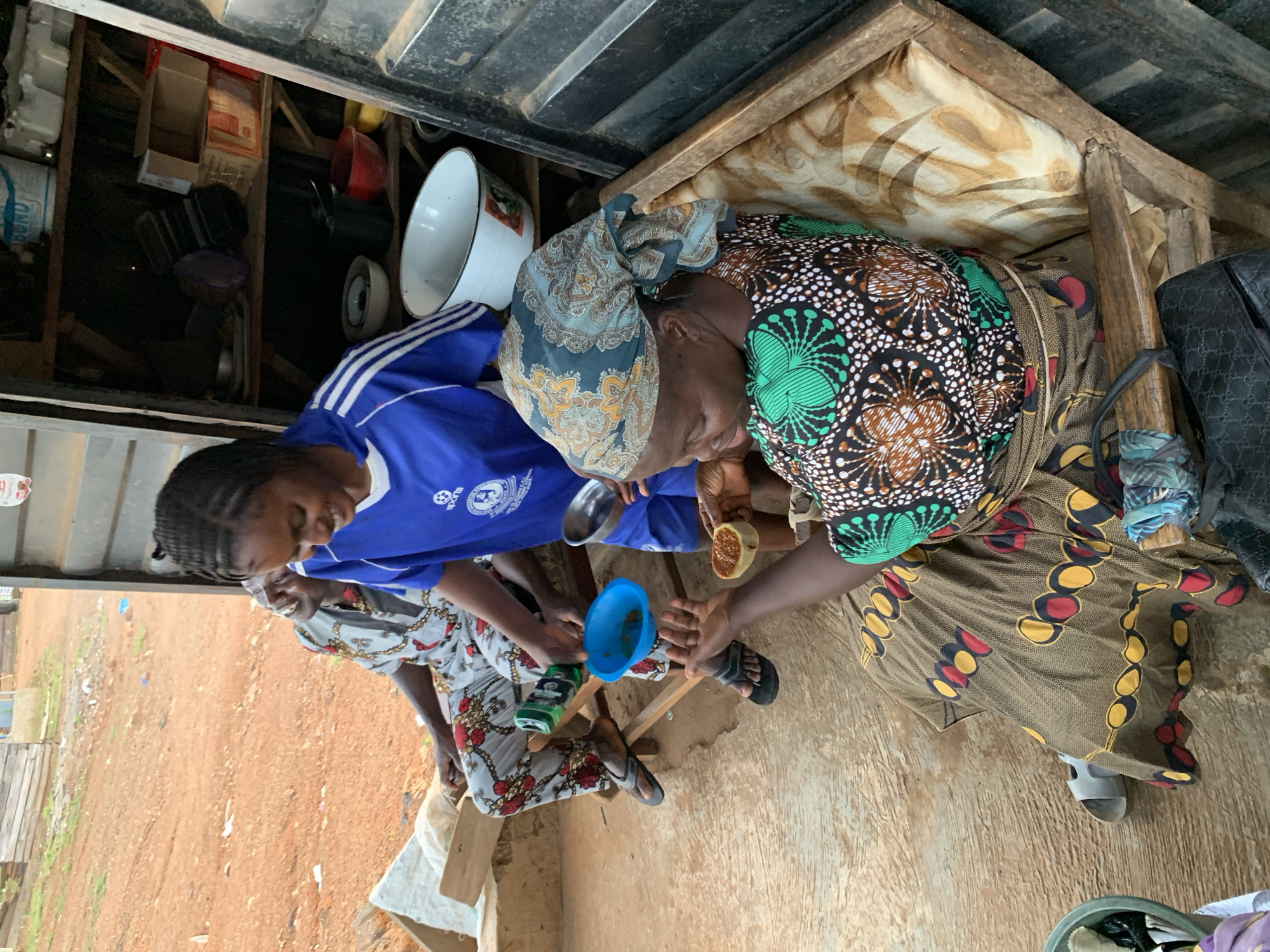 Three women are seated outdoors in Kabba, Nigeria, apparently processing food. One woman, wearing a blue soccer jersey, sits to the left, smiling, while operating a small blue plastic funnel. Another woman is facing away from the camera, wearing a colorful print headscarf and brown and yellow dress, holding a piece of fruit in her hand. The third woman, seated at the bottom left corner, is in a floral print top. They sit on a wooden frame. Behind the woman in blue is a wooden shelf with kitchen items and a white enamel bowl. The ground is brown earth and there is a corrugated iron roof overhead.