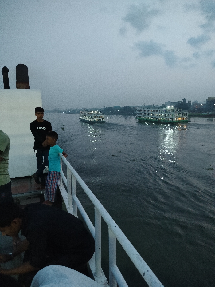 The image shows a view from the deck of a ferry, looking out over the Buriganga River in Zazira, Bangladesh. The ferry is moving forward. There are four people visible on the deck, with a white railing on the right side of the image. The ferry is heading towards the left of the image, with a large passenger ferry on the left, and another larger passenger ferry on the right. The sky is overcast, the water is choppy and dark, and the sun is setting in the background. The image was taken in the evening, just before sunset. The image appears to have been taken with a smartphone.