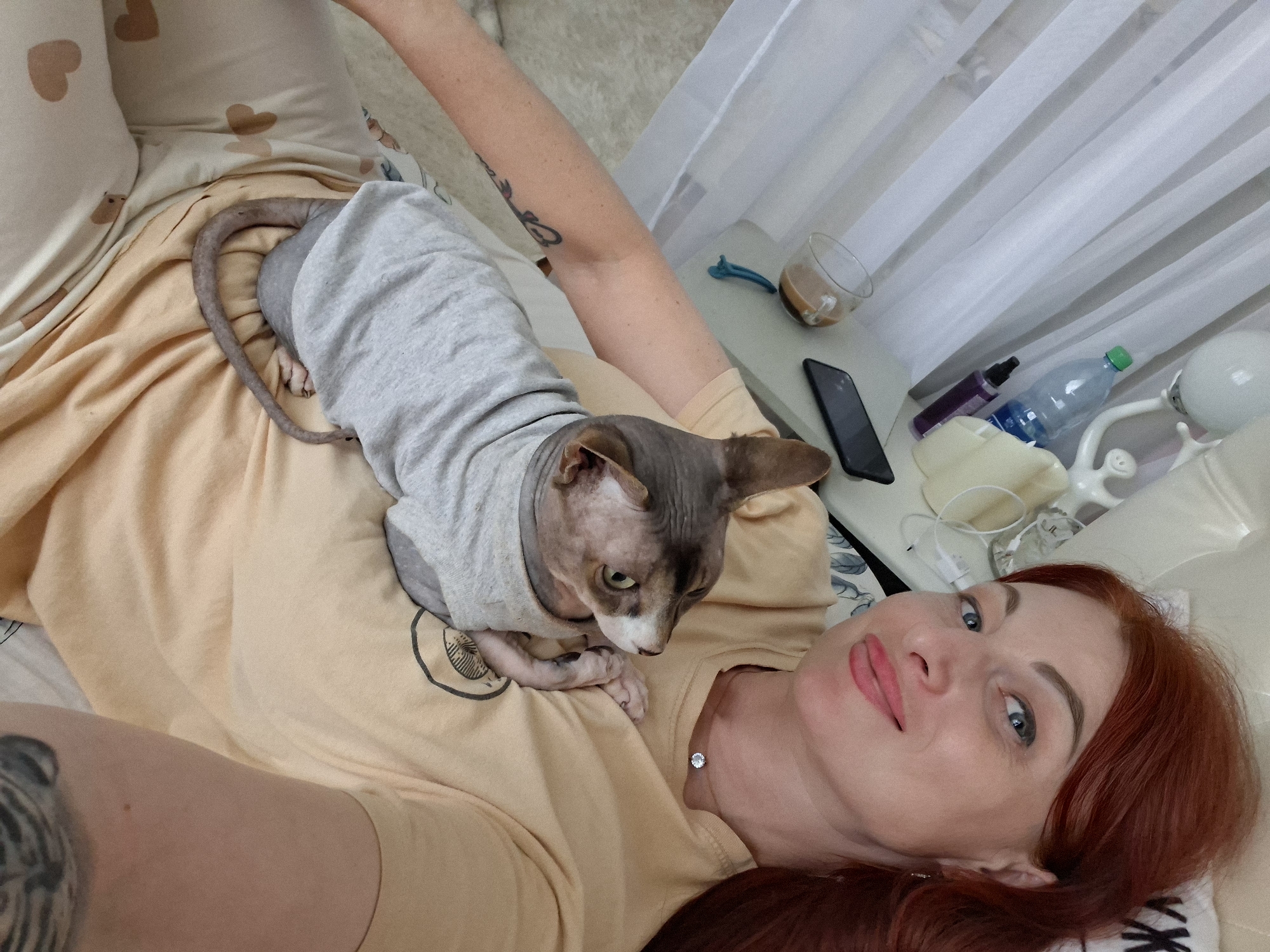 A woman with long, reddish hair and blue eyes is lying in bed, holding a Sphynx cat. She is wearing a light yellow t-shirt and light-colored pajama bottoms with a heart pattern. The cat is wearing a grey t-shirt. A bedside table is visible with a glass of coffee, a bottle of water, a phone, and a lamp. The scene appears to be indoors, likely in a bedroom. The provided metadata indicates the location as Myadzyel, Belarus.