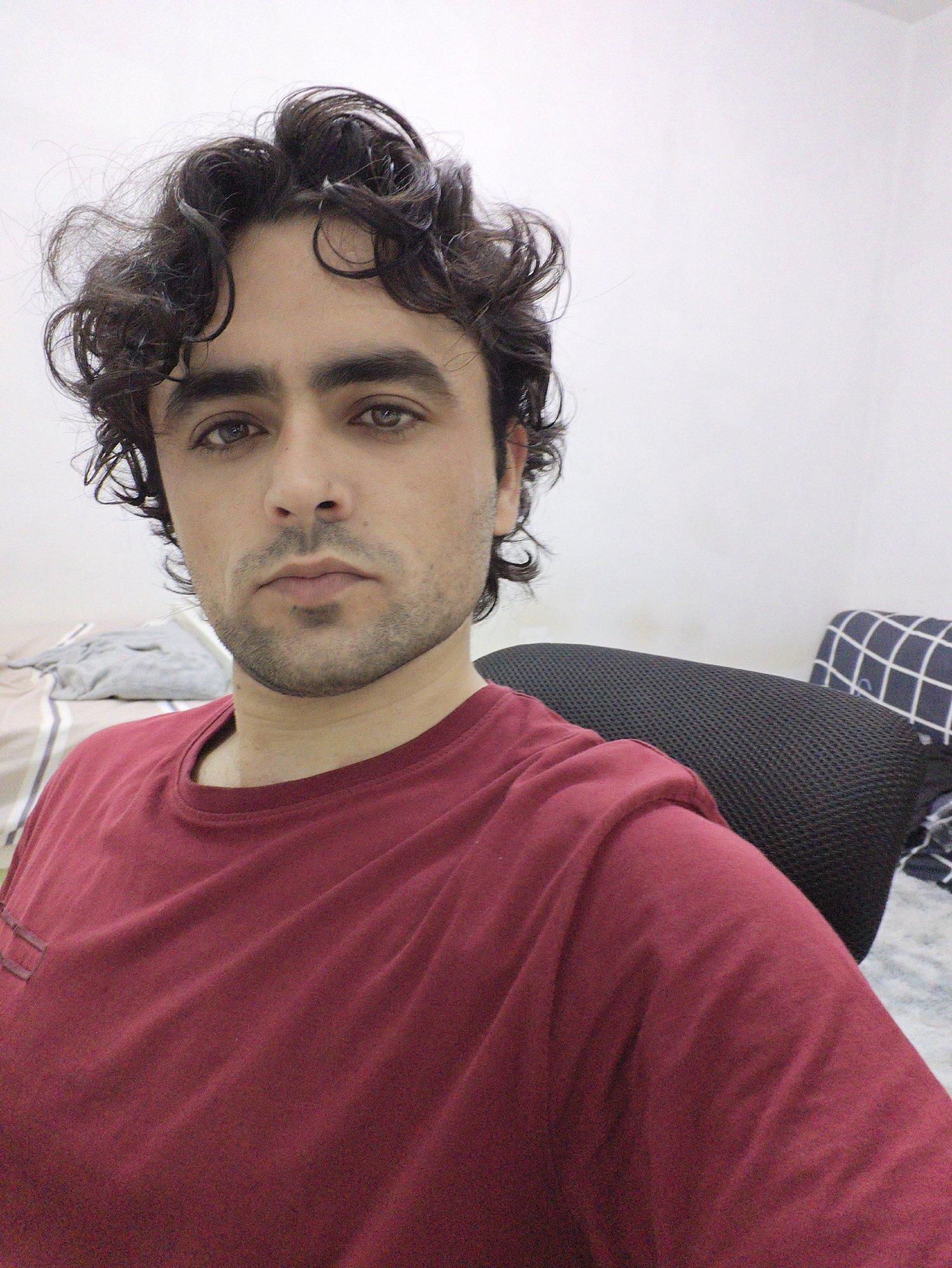 A person is shown in a self-taken photograph. The individual is male, with dark curly hair and facial hair. He wears a maroon t-shirt and is sitting on a black office chair. The background consists of a white wall and a bed with striped sheets and a pillow with a grid pattern. The location is not discernable and cannot be determined to be in the Chongqing High-tech Zone, China, based on the image alone.