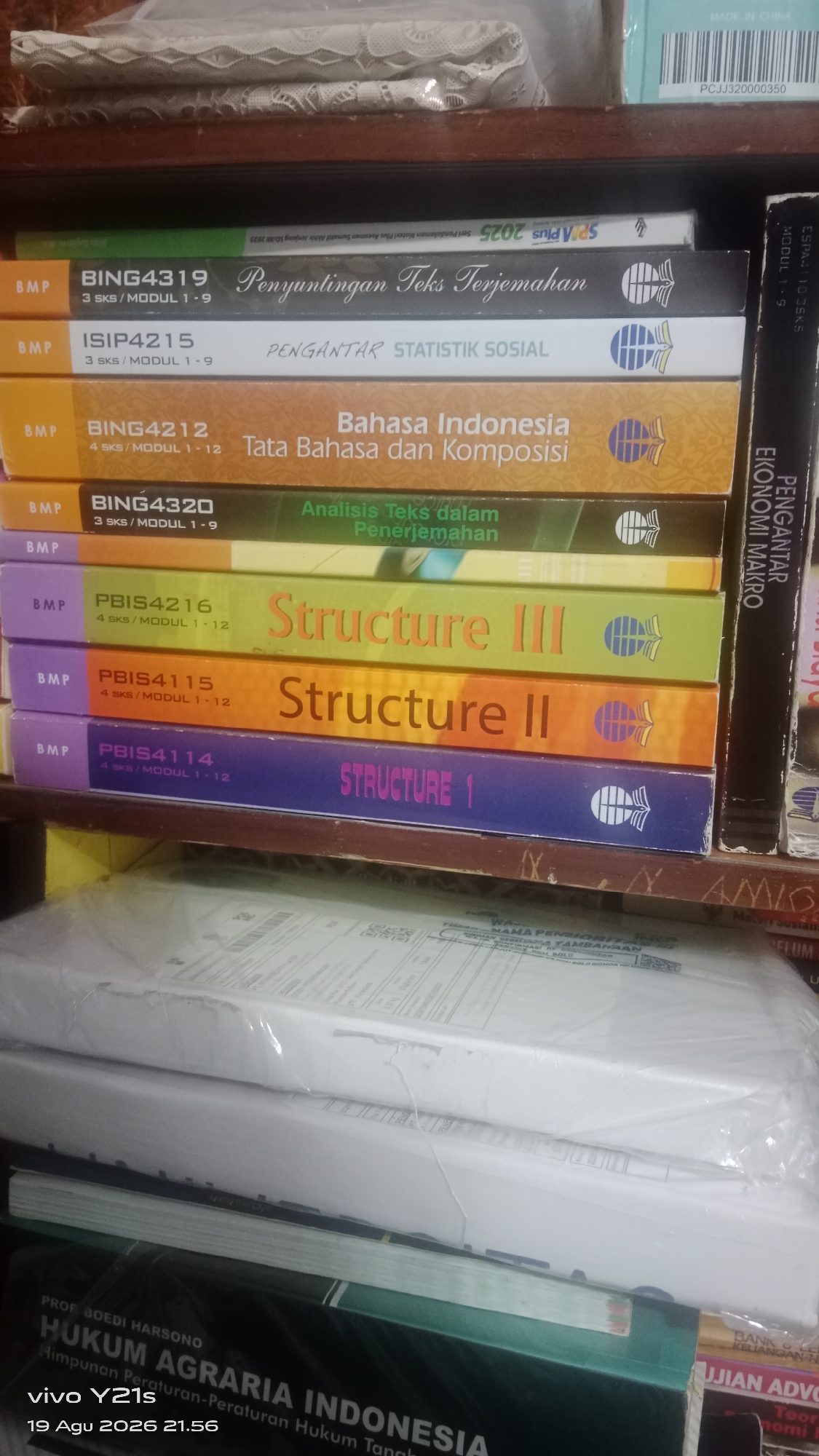 The image shows a bookshelf filled with books. The books are stacked horizontally and vertically. Some of the books have colorful spines with titles and author names. In the foreground, there are two large packages wrapped in plastic. The bottom of the image shows a book with the title "HUKUM AGRARIA INDONESIA". There are no people visible in the image. The environment appears to be indoors, likely a room with a bookshelf.