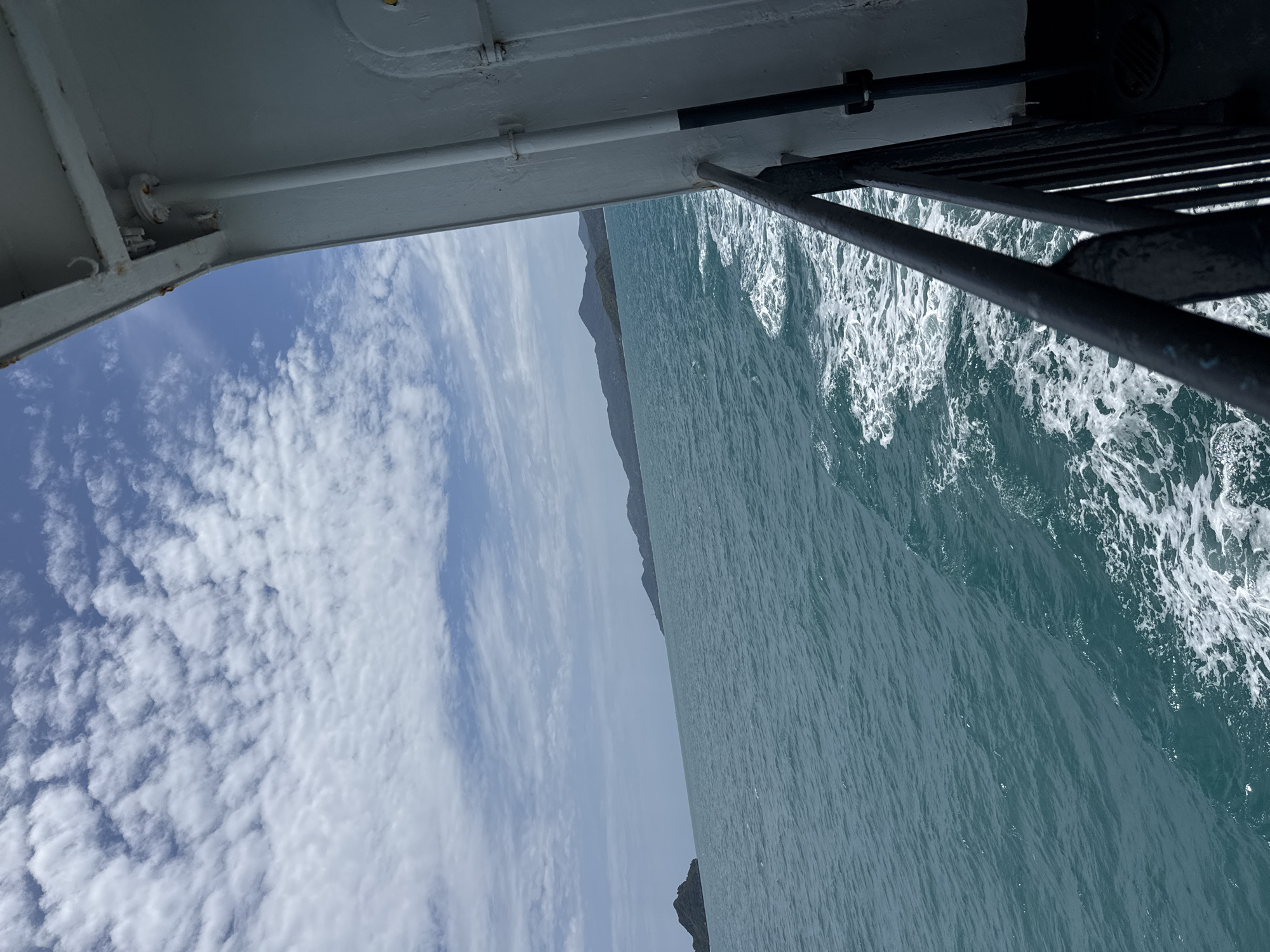 The image depicts a view from the side of a ferry boat traveling through turquoise ocean water. The ferry's white hull and a black metal railing are visible on the right side of the frame. The water is choppy, with white foam trailing behind the boat. In the distance, several islands covered in lush green vegetation rise from the sea. The sky is a clear blue, dotted with scattered white cumulus clouds. The scene suggests travel between islands, a common mode of transportation in Ko Pha-ngan, Thailand, known for its numerous islands and coastal activities.