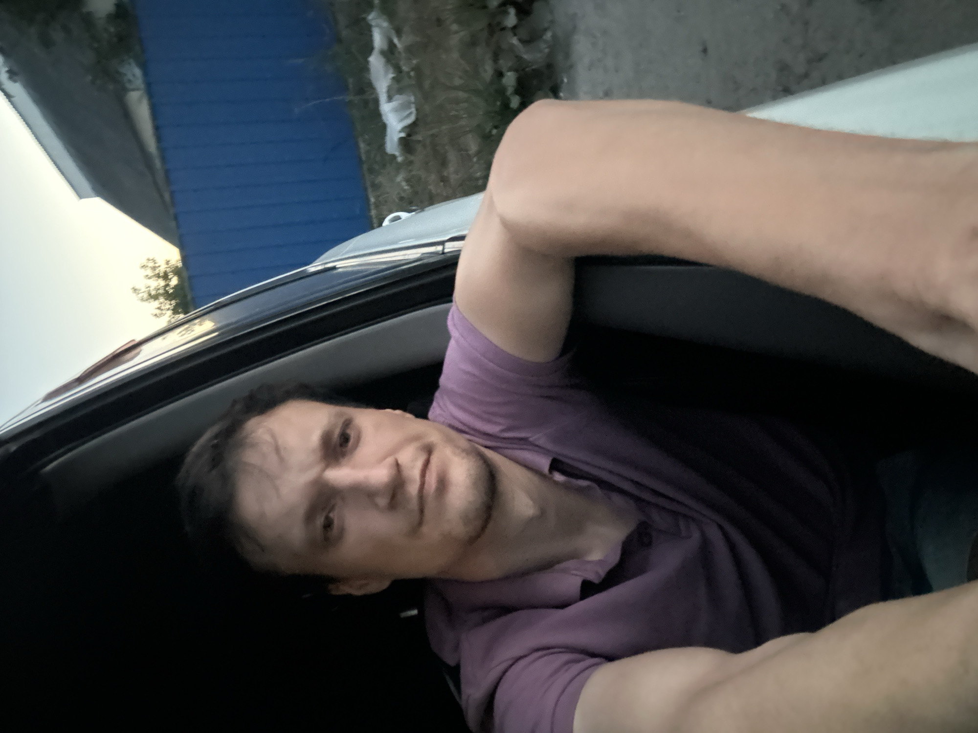 A medium shot captures a man sitting in the driver's seat of a car, looking towards the camera with a slight frown. His right arm rests on the open car door, revealing a portion of his tanned skin. He is wearing a short-sleeved purple polo shirt. In the background, a blue corrugated metal fence stretches across the frame, separating a yard with dry grass and scattered white debris from a building with a dark roof and a white overhang. The sky above is a pale blue, suggesting either early morning or late evening. The scene is set in the city of Shatava, Ukraine.