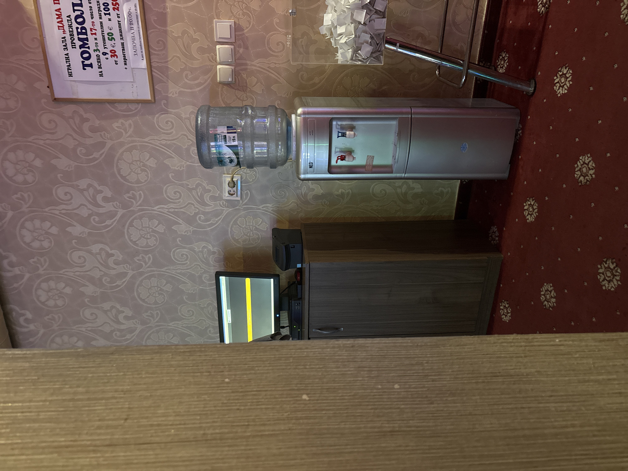 The image depicts the interior of a room, likely a gaming hall or reception area, in Karlovo, Bulgaria. A sign on the wall, written in Bulgarian, advertises a "TOMBOLÁ" (lottery or raffle) organized by "IGRALNA ZALA 'DAMA PO'" (Gaming Hall 'Lady Of'). The sign details prize amounts in Euros, with winning tickets drawn on the 3rd and 17th of each month, and a jackpot prize of 250 Euros. It also states that the lottery starts at 20:00.
In the foreground, a light brown wooden partition is visible on the left. To the right of the partition, a grey cabinet stands against a wall adorned with a patterned wallpaper. On top of the cabinet, a computer monitor displays a yellow and grey interface, and a black printer is positioned next to it.
A silver water cooler with a clear plastic water jug on top is placed in the center of the image. The water cooler has red and blue dispensing taps. To the right of the water cooler, a clear plastic container filled with numerous small white paper slips is visible, suggesting it's a collection box for lottery entries. A metal bar stool with a chrome finish stands nearby. The floor is covered with a red carpet featuring a repeating floral pattern. The lighting appears to be artificial, casting a soft glow on the scene. There are no people visible in the image.
