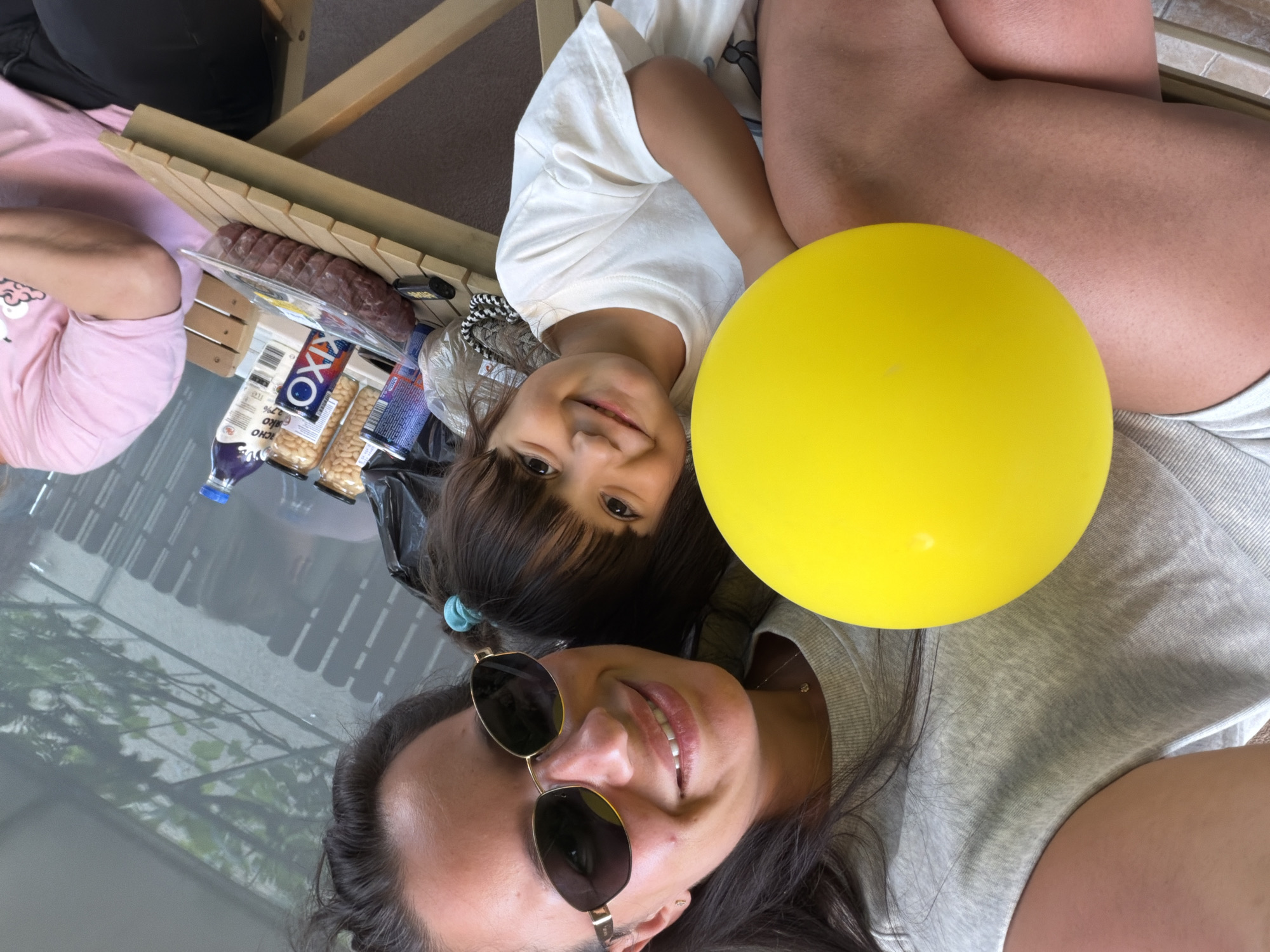 In the city of Burgas, Bulgaria, a close-up, slightly tilted selfie captures a woman and a young child. The woman, wearing a grey t-shirt and sunglasses, is positioned in the upper left, her arm around the child who is held in her lap. The child, in a white t-shirt, faces forward and smiles. A large, bright yellow balloon dominates the left side of the frame, partially obscuring the child's body and the woman's arm. In the background, a wooden table is laden with various items, including jars of food, cans, and a plastic container of raw meat. A pink-clad individual is partially visible on the right side of the image. The setting appears to be an outdoor area, possibly a backyard or patio, with a blurred fence and foliage visible behind the table.