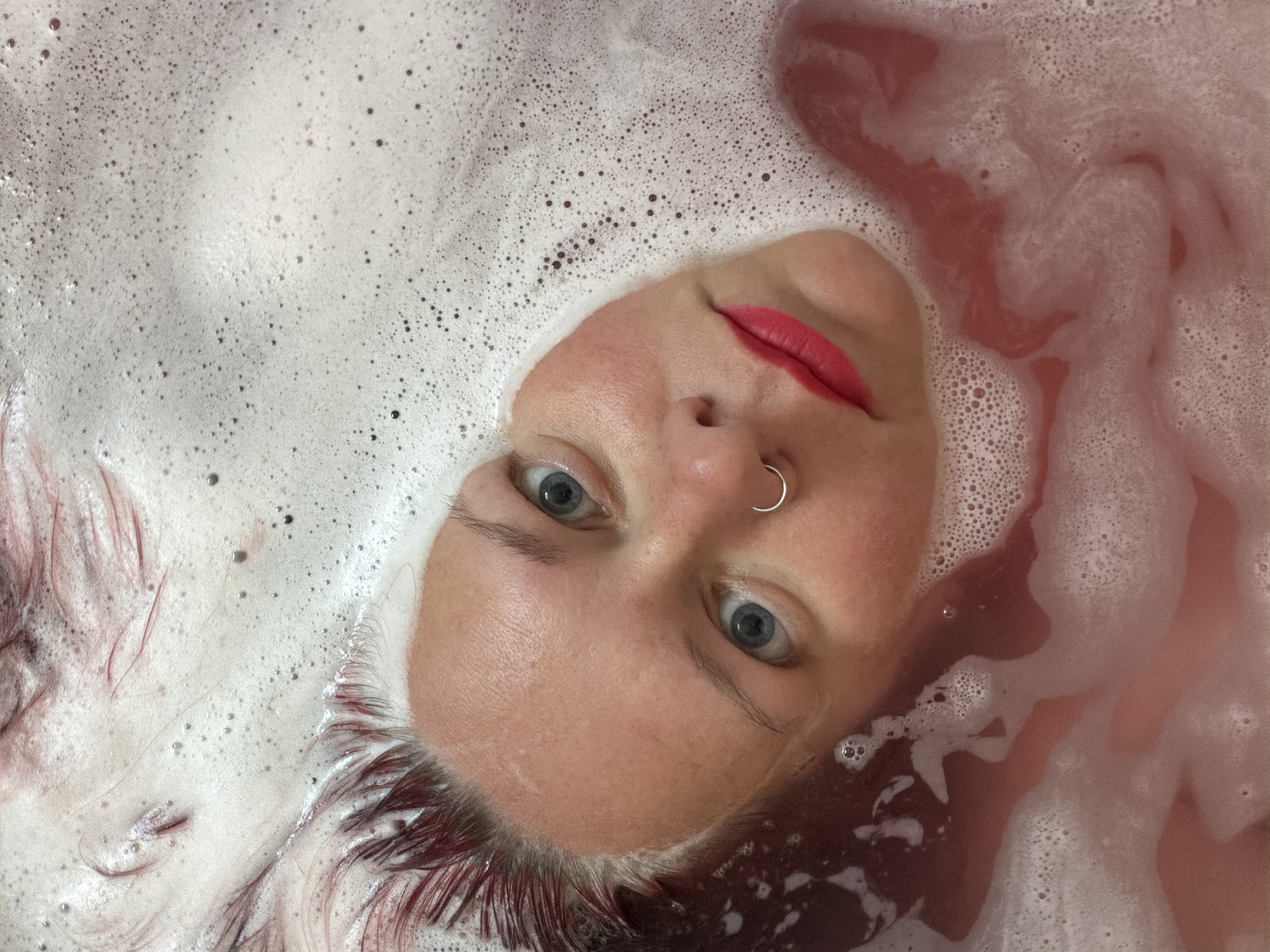 A woman's face is visible amidst water and bubbles. The woman has fair skin, blue eyes, a nose ring, and red lipstick. Her hair appears to be red/purple and wet. She is surrounded by white soap bubbles and reddish-tinted water. The image is shot from a close, slightly downward angle, giving the impression that the woman is submerged in the water. Location: Unknown, Unknown.