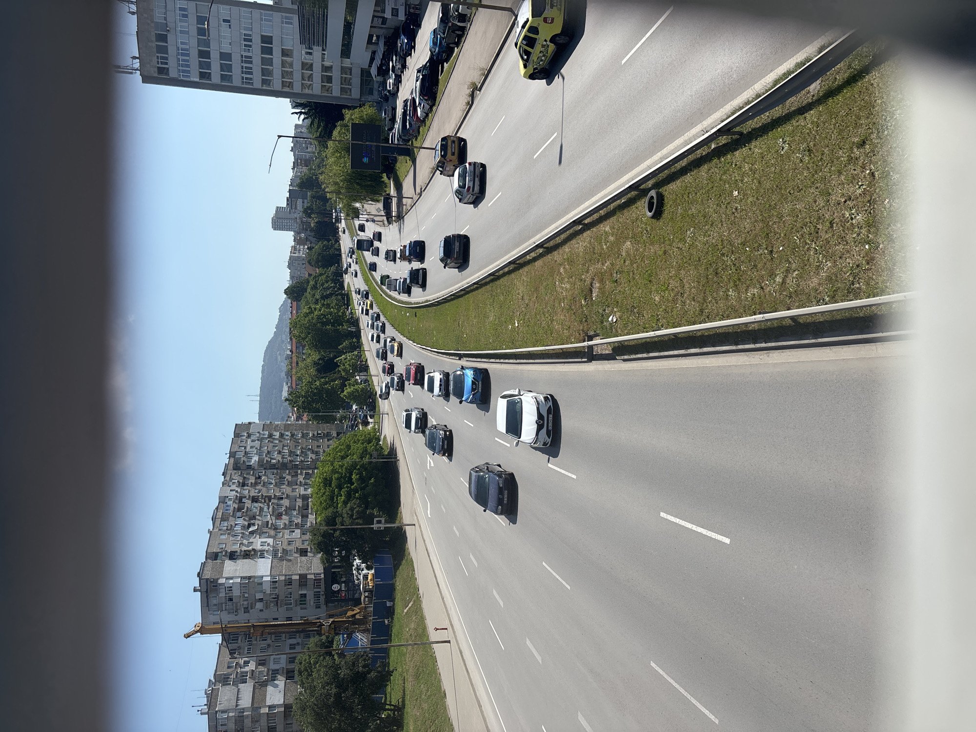 The image captures a multi-lane highway in Varna, Bulgaria, viewed from an elevated perspective, possibly a bridge or overpass. The highway is busy with traffic, with numerous cars and a bus visible. The road is paved and marked with white lane lines. On the left side of the highway, there are apartment buildings and a construction crane. On the right side, there is a grassy median with a metal guardrail, and beyond that, more buildings, including a tall, modern office building. The sky is clear and blue, indicating a sunny day. The overall scene depicts an urban environment with significant vehicular activity.