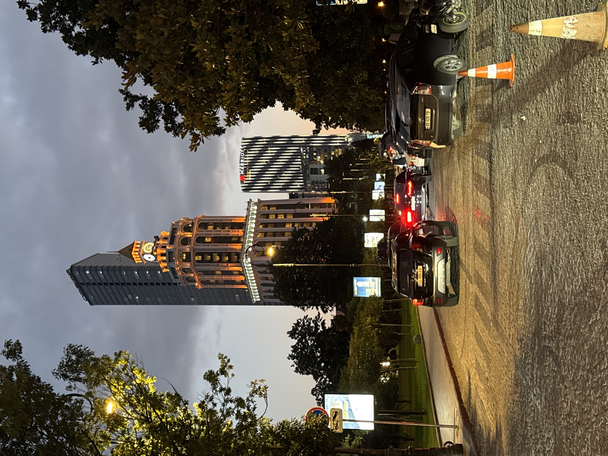 The image depicts a street scene in Batumi, Georgia, at dusk. A tall, illuminated building with a clock tower is prominently featured, casting a warm glow against the twilight sky. Adjacent to it, another modern skyscraper with a striped facade, identified by a Marriott logo, stands tall. The street is paved with cobblestones and lined with trees. Several cars are visible, some with their brake lights illuminated, suggesting they are stopped or moving slowly. Traffic cones are placed on the road, indicating potential construction or a designated area. In the background, streetlights and illuminated signs contribute to the urban ambiance. The overall scene conveys a sense of a city transitioning from day to night, with architectural landmarks and vehicular traffic.