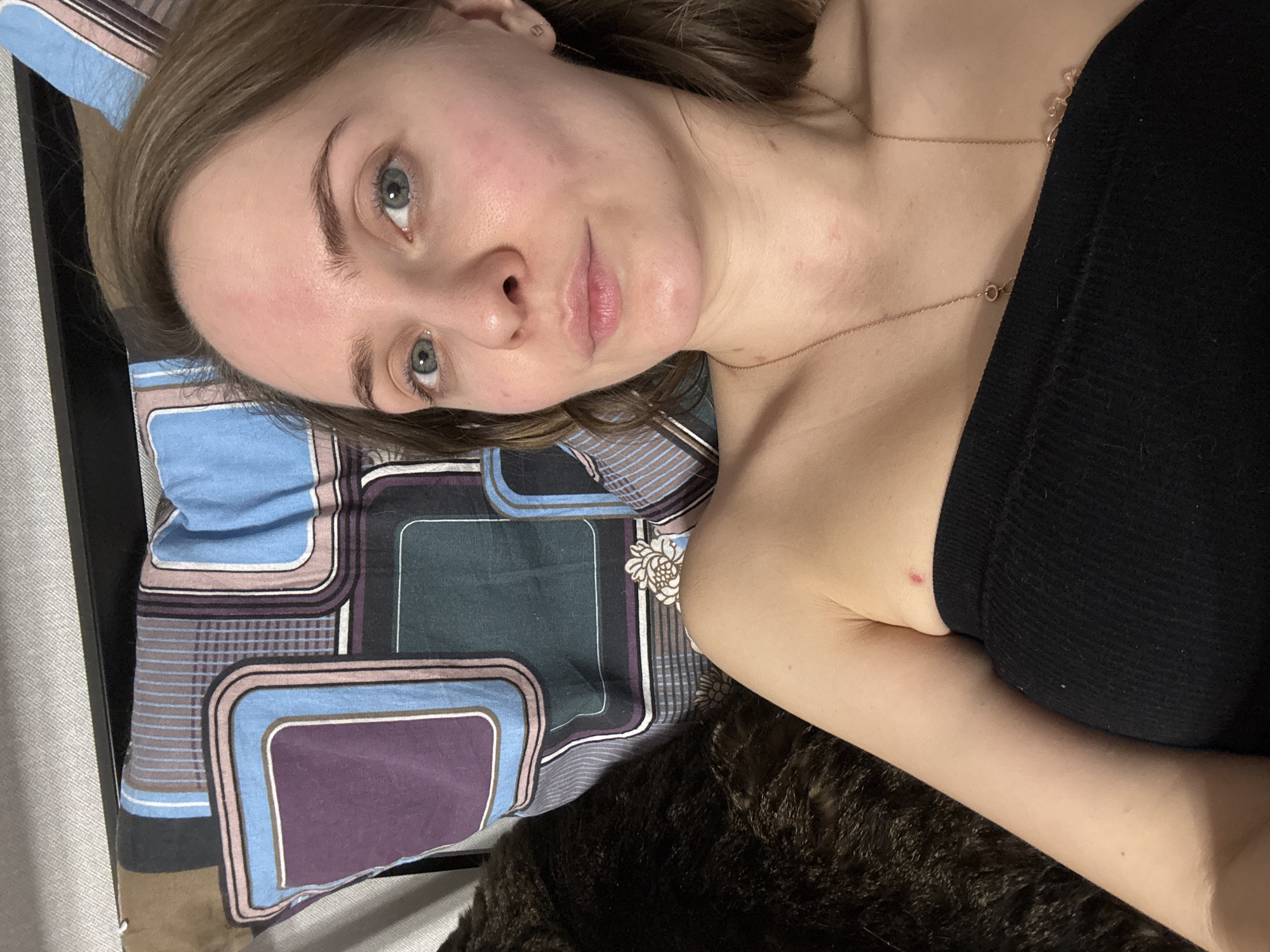 A fair-skinned female with shoulder-length brown hair is lying down on a bed. She has light blue eyes, a straight nose, and slightly parted lips. She is wearing a black strapless top and a gold necklace. The bed has a pillow with a geometric pattern in shades of blue, purple, and brown. A dark, furry object, possibly a pet, is also on the bed near the woman. The background is out of focus, but a wall is visible in the upper left corner. The location is not discernible from the image.