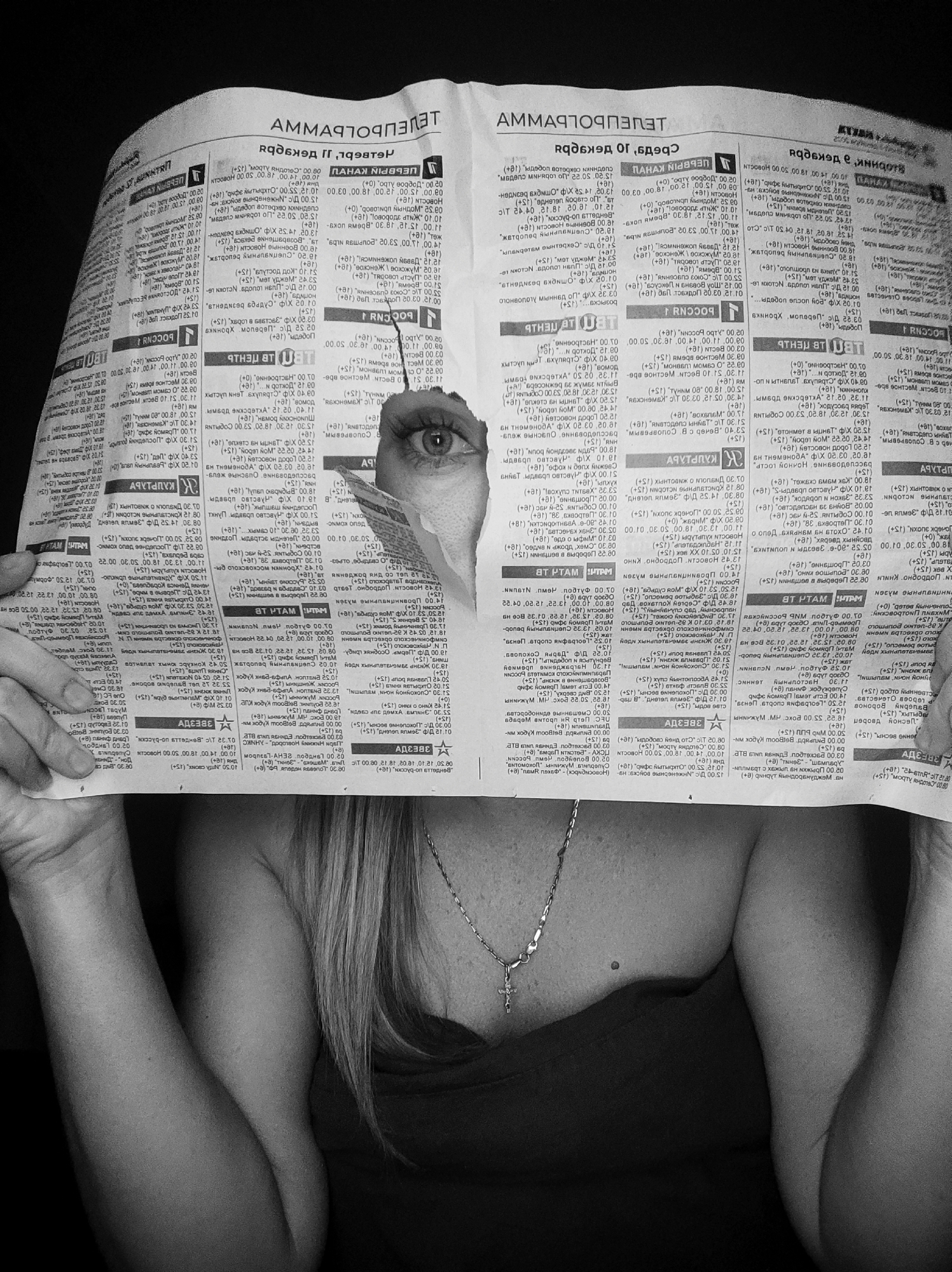 This black and white portrait features a person, likely female, partially obscured by a newspaper held up to their face. One eye of the person is visible, peeking through a roughly torn hole in the newspaper and looking directly at the viewer. Below the newspaper, a shoulder, arm, and chest are visible, along with long, light-colored hair (appearing grey/blonde in monochrome). A dark garment covers the chest, and a silver-colored chain with a crucifix pendant adorns the neck.
The newspaper is covered in Cyrillic text, confirming its origin in Russia, consistent with the Votkinsk location. The main title, "ТЕЛЕПРОГРАММА" (TV Program), is visible but appears mirrored in the original image. Other mirrored text includes channel names like "РОССИЯ 1" (Russia 1), "ПЯТЫЙ КАНАЛ" (Fifth Channel), and "НТВ", along with television program listings, various time slots (e.g., 00.00, 06.00, 12.00), and age ratings such as "(+0)", "(+6)", "(+12)", "(+16)", and "(+18)". The word "СРЕДА" (Wednesday) is also discernible, likely indicating the day of the TV schedule. The activity portrays a sense of hidden observation or playfulness. The setting appears to be indoors, with a dark, indistinct background.