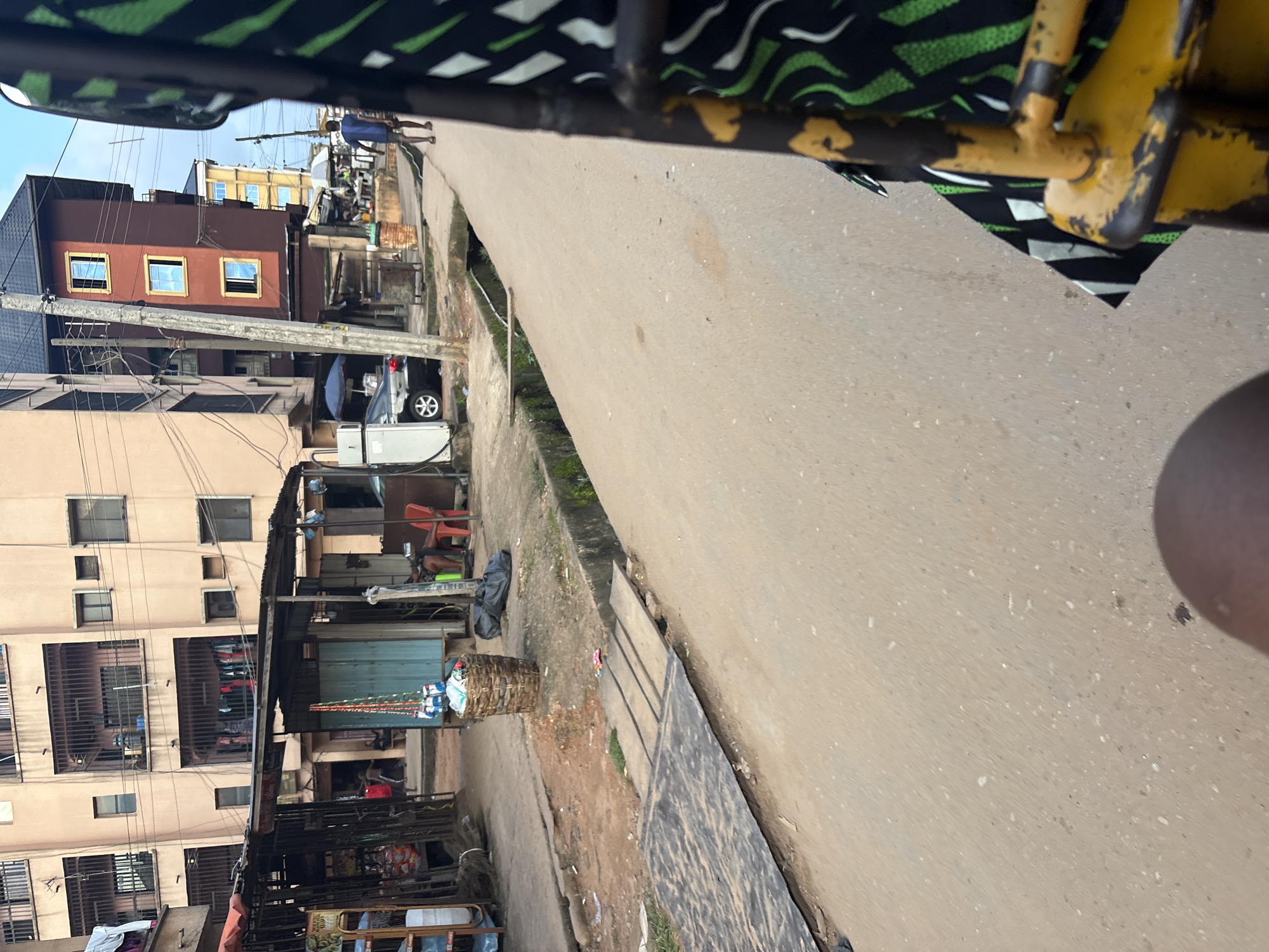 The image captures a street scene in Onitsha, Nigeria, likely from the perspective of a passenger in a vehicle. In the foreground, the edge of a yellow metal frame and a patterned fabric, black and green, are visible. The street itself is paved with a light brown, slightly textured surface, with some debris and scattered white specks. A narrow, open drainage channel runs alongside the road, covered in places by wooden planks.
To the left of the road, a series of informal market stalls are set up. One stall has a corrugated metal roof and displays various goods, including bottles and what appear to be woven baskets. In front of a stall, a person is seated on a red plastic chair, partially obscured. A large, woven basket filled with items sits near the drainage. Further back, a multi-story building with balconies and barred windows dominates the background. Clothing is seen hanging on lines in front of some of the balconies. Another building, painted a deep red with yellow-trimmed windows, stands to the right of the first building. A dark-colored car is parked near a white meter on the left side of the street. A man in a blue shirt walks away from the camera down the street. The sky above is a pale blue with some wispy clouds. The overall impression is of a bustling, somewhat informal urban environment.