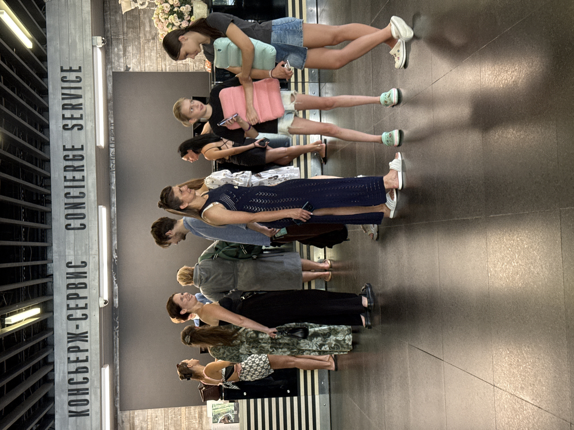 The image shows a group of people standing in what appears to be a hotel lobby or reception area. Above them, a sign reads "КОНСЬЕРЖ-СЕРВИС CONCIERGE SERVICE" in large, bold letters. To the left, a woman in a patterned dress and a woman in a black jumpsuit are standing. In the center, a woman in a dark blue dress is holding a phone. To her right, a young girl in shorts and a black t-shirt is holding a pink inflatable pillow, and another girl in a denim skirt and t-shirt is holding a light blue inflatable pillow. In the background, there are more people, including a man in a blue shirt and a woman in a grey robe. The environment appears to be indoors, with a dark tiled floor and a ceiling with exposed beams and lighting fixtures. The overall scene suggests a public space where people are waiting or interacting. There is no information in the image to suggest the location is in the city of Yalta, Ukraine.