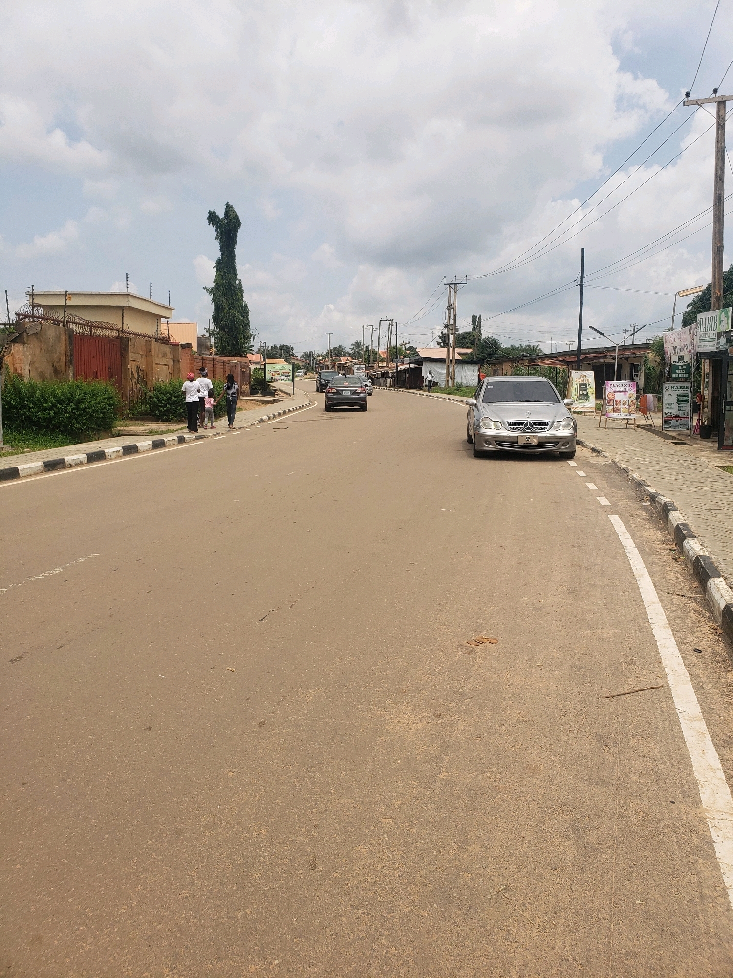 The image shows a street scene in Narayi, Nigeria, under a cloudy sky. A paved road with white dividing lines runs through the center of the frame, curving slightly to the right. On the left side of the road, a brown fence with wire topping encloses a property with a building. A tall, slender tree with dense foliage stands behind the fence. Several people, including two adults and a child, are walking on the sidewalk on the left side of the road.
On the right side of the road, a sidewalk runs alongside several businesses with awnings and signs. A silver Mercedes-Benz sedan is parked on the right side of the road, facing the camera. Several other cars are visible in the distance on the road, and more buildings and trees line the street further back. A utility pole with wires is also prominent on the right side. The ground on the road shows some scattered debris.
