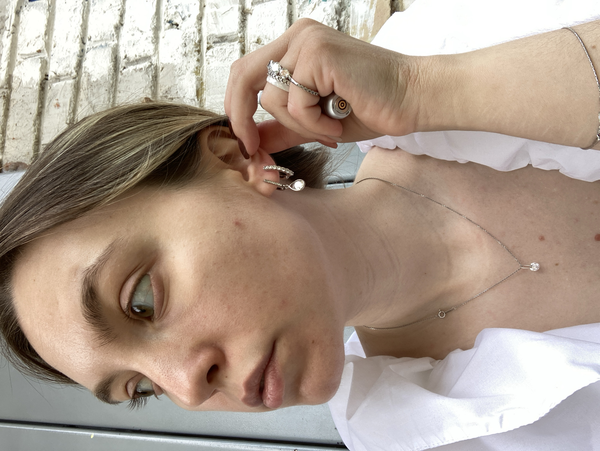 A woman with fair skin, light brown hair with blonde highlights, and light brown eyes is seen in a medium shot. She wears a white, ruffled top and jewelry including a silver necklace with a single diamond, a silver bracelet, and several rings on her right hand. The woman is adjusting a silver ear cuff with a teardrop-shaped pearl on her right ear. The background shows a brick wall. There is no information available about the location in the city of Chaikovsky, Russia.