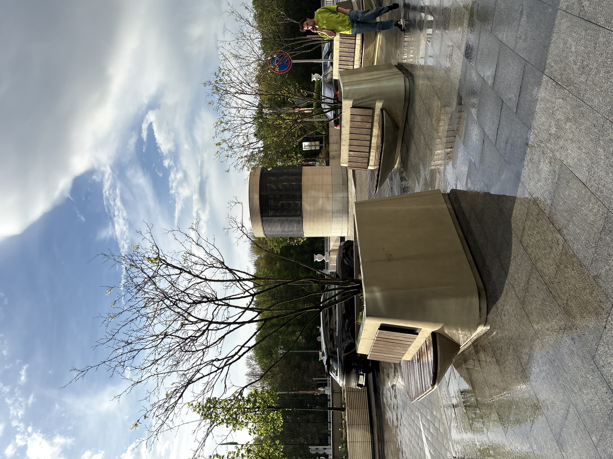 The image is of a street in Minsk, Belarus. The scene is wet, likely after a recent rain, and the ground reflects the sky and surrounding objects. Several modern concrete benches with wooden slats are placed along the street. A young man in a bright green shirt and blue jeans is visible in the background, standing and talking on a cell phone. A car is parked alongside one of the benches. Trees are planted within the benches. A cylindrical building with a striped facade is visible in the mid-ground. A no-parking sign is present. The sky is cloudy with patches of blue.