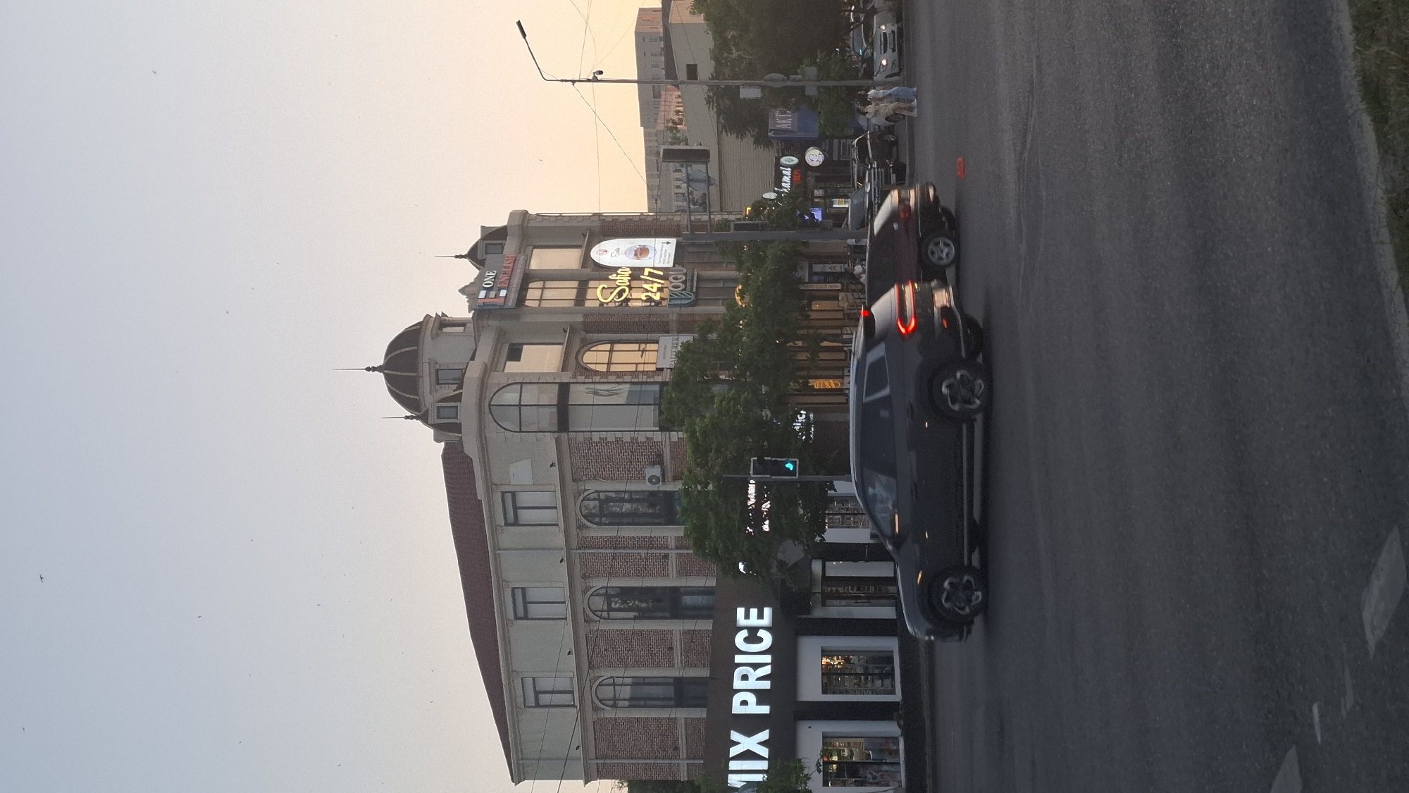 The image shows a street scene in Shymkent, Kazakhstan, likely during the late afternoon or early evening as indicated by the warm, soft lighting. A multi-story building with a reddish-brown tiled roof and a domed turret dominates the left side of the frame. The building features a mix of brick and stucco facades, with arched windows and balconies. Visible signage on the building includes "ONE ENGLISH," "Salon 24/7," and "MIX PRICE." A dark-colored car with its brake lights illuminated is in the foreground, on the right side of the street, suggesting it is either stopped or moving slowly. Another dark car is visible further down the street. A traffic light with a green signal is also present. The sky is a pale, hazy color, typical of dusk or a slightly overcast day. The overall impression is a snapshot of everyday urban life in Kazakhstan.