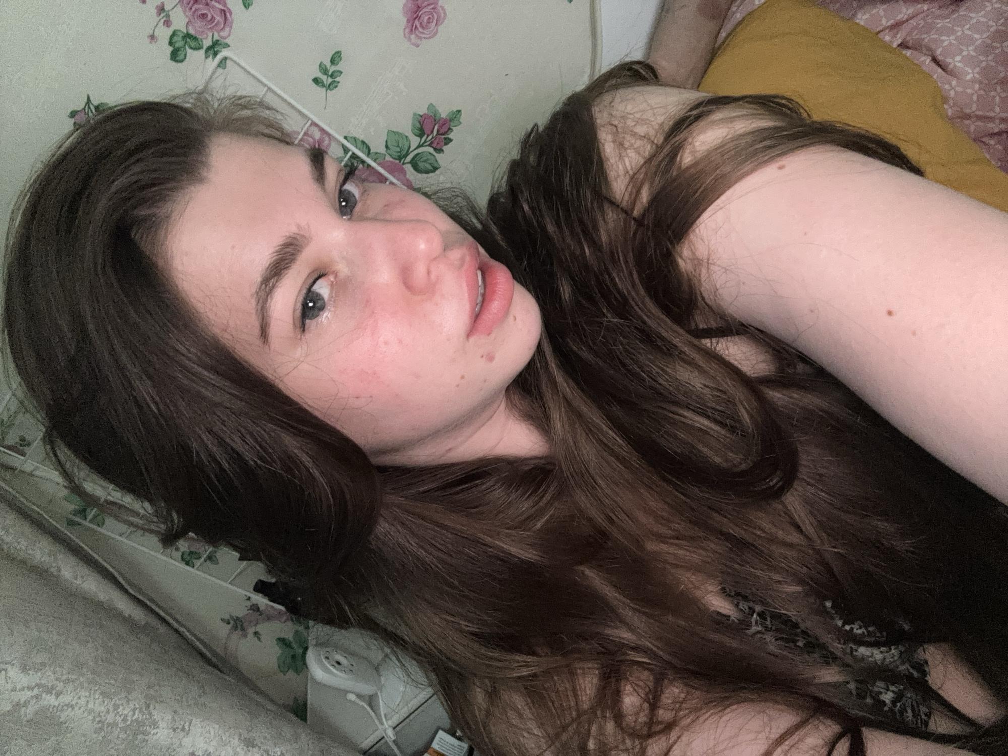 A young woman with long, dark brown hair is lying down, looking at the camera. She has fair skin and gray eyes. Her head rests on a pillow with a floral pattern. A yellow cushion or blanket is visible behind her. An old-fashioned white telephone is situated near the head of the bed. It is impossible to ascertain a location in the city of Мещёры, Russia, from the image provided.