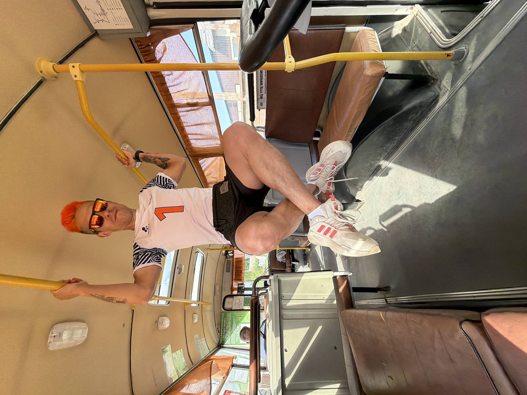 A man with orange hair and sunglasses is hanging upside down in a bus or tram. He is wearing a white T-shirt with the number "1" on it, black shorts, white socks, and white Adidas sneakers with red stripes. He is holding onto yellow handrails with both hands and has his legs crossed. The bus or tram has brown leather seats and yellow handrails. The windows show a view of buildings and trees. The location is Chaikovsky, Russia, based on contextual cues that the image originates from.