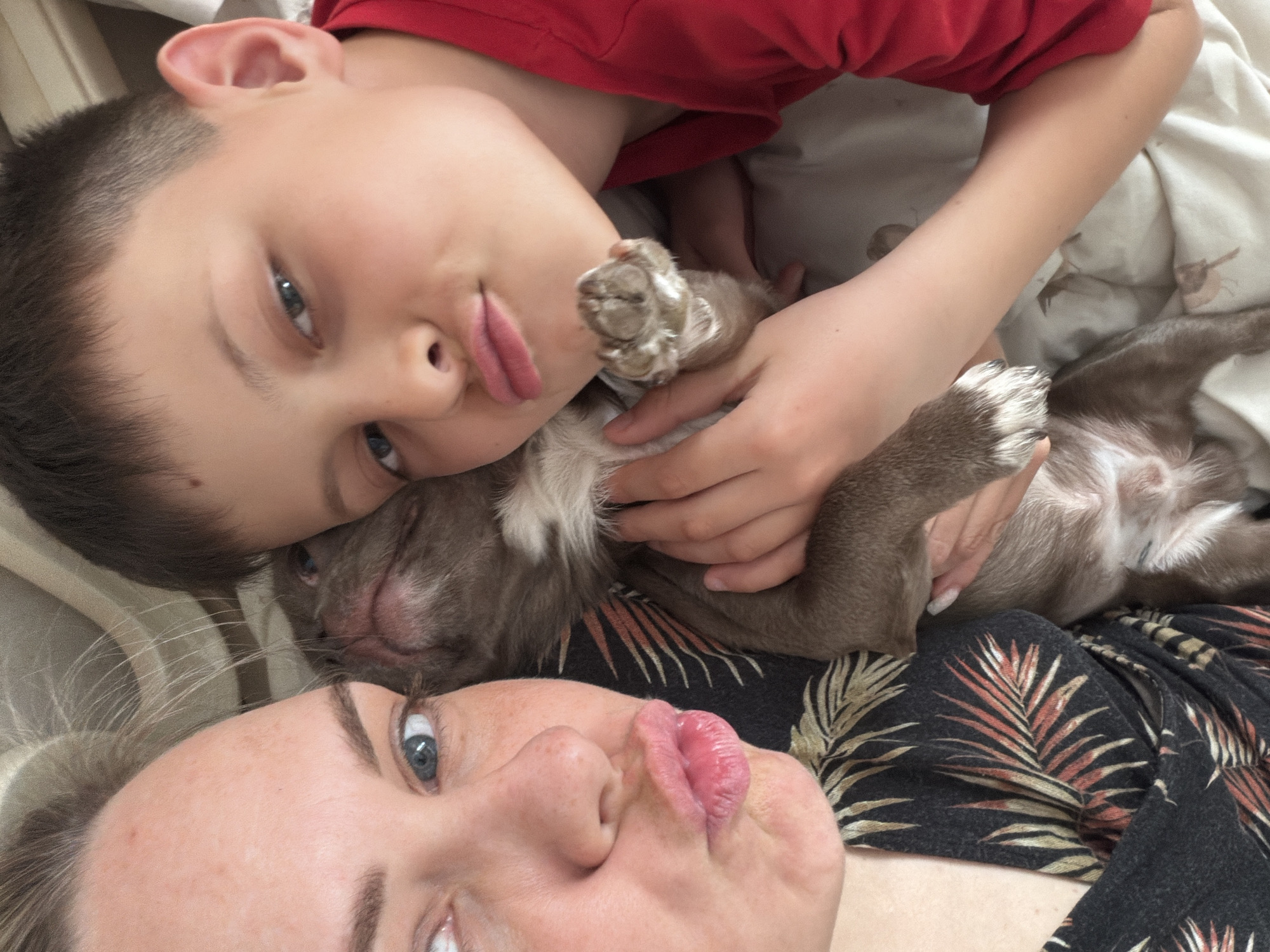 A close-up, slightly tilted shot depicts a fair-skinned woman and a young boy, both facing the camera with pursed lips. Sandwiched between them is a small, brown and white puppy held in the boy's hands. The boy is wearing a red shirt, while the woman is wearing a black top with a pattern of brown and white palm leaves. The location is indiscernible, and there is no indication of Votkinsk, Russia.