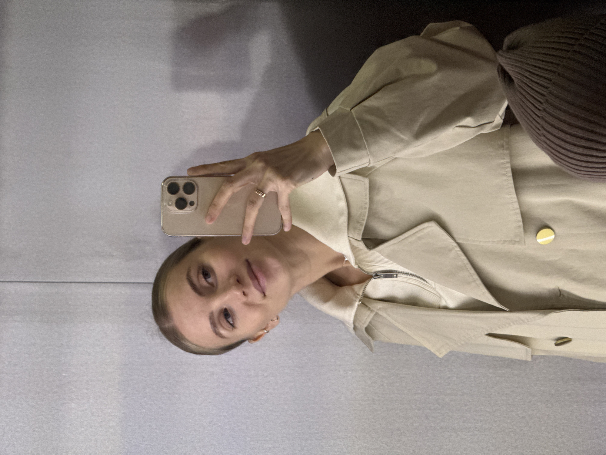 A woman takes a selfie indoors, holding a gold-colored smartphone in her left hand. She is wearing a light beige trench coat and a brown knit hat is partially visible near her shoulder. She has fair skin, blonde hair, and is wearing a ring on her left ring finger. The background appears to be a gray or lightly colored wall. The location is unknown and is not explicitly identifiable as Izhevsk, Russia.