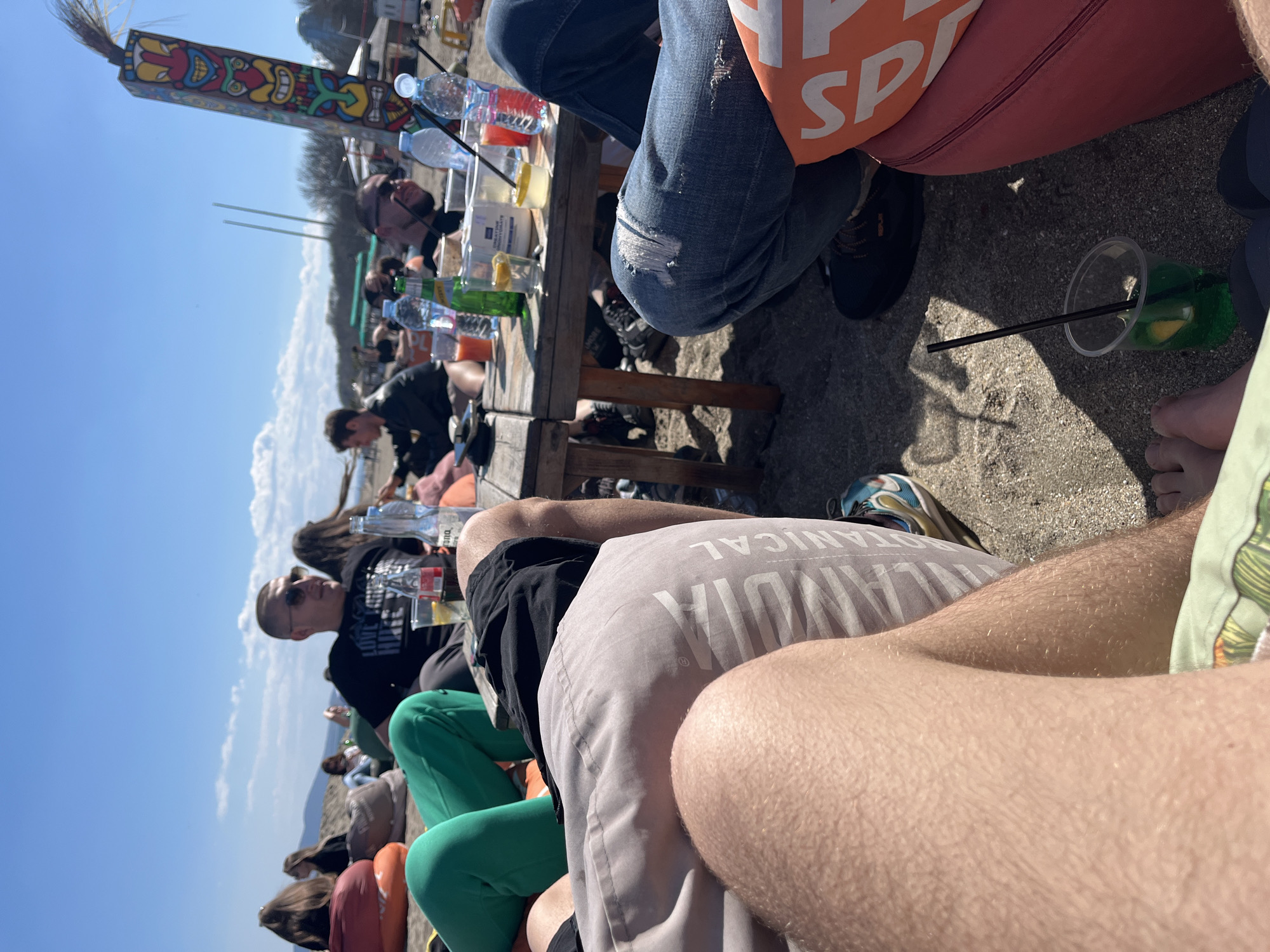 This outdoor scene depicts a group of people relaxing at a beachside establishment in Burgas, Bulgaria, on a sunny day. The perspective suggests the photo was taken from the vantage point of someone seated among the group. A wooden table laden with various beverages, including water bottles and mixed drinks, sits in the center, indicating a social gathering. The table is surrounded by individuals seated on beanbag chairs. The focus is on the people, particularly their clothing and posture, which suggest a casual and relaxed atmosphere. In the background, the beach and waterfront are visible under a clear blue sky. A colorful tiki-style structure stands out against the sky, adding a distinct cultural element to the setting.