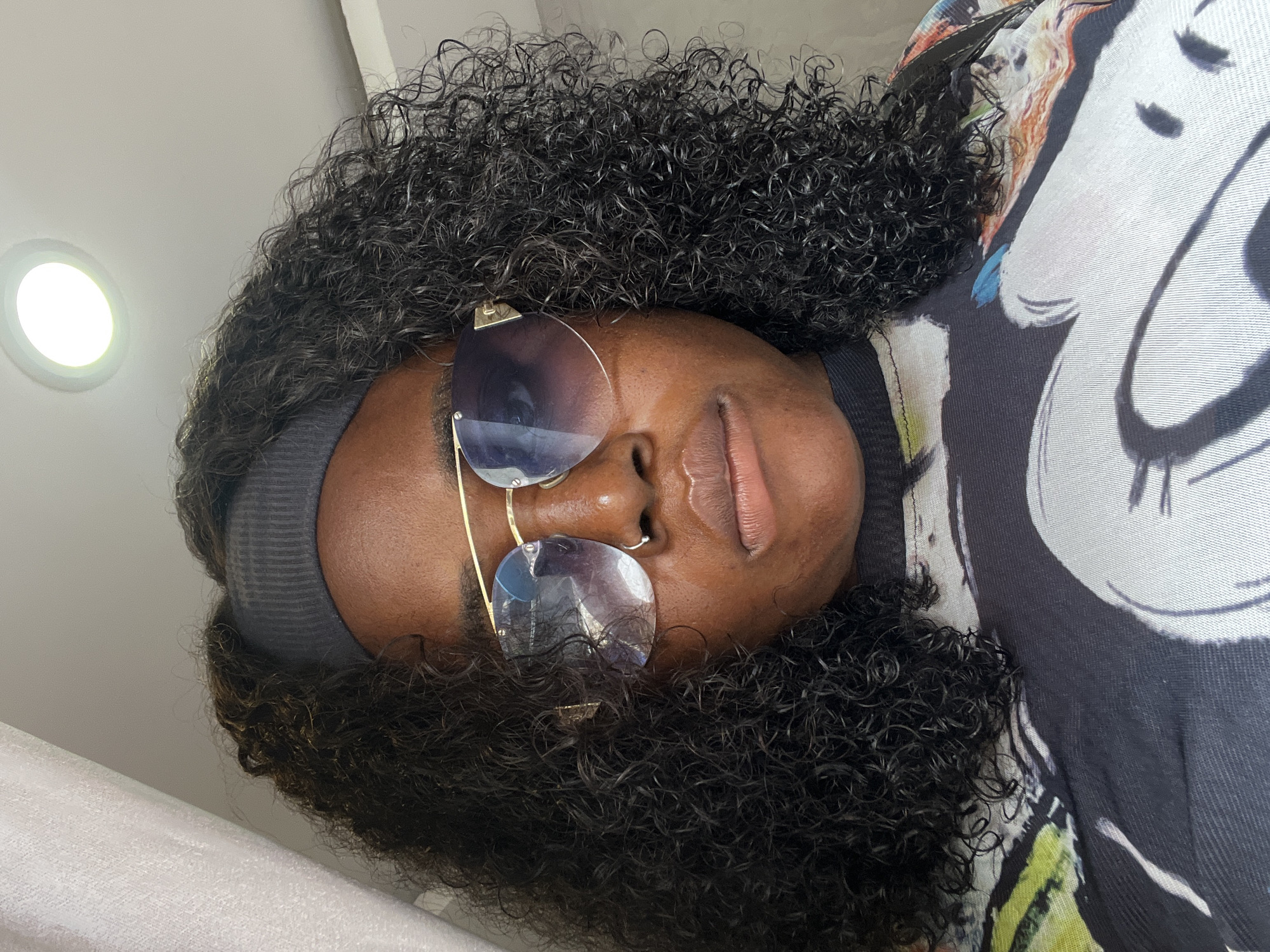 A medium-shot, eye-level selfie shows a person with dark, curly hair and brown skin, wearing large, gold-rimmed sunglasses with blue lenses and a grey headband. They are also wearing a black t-shirt with a multi-colored abstract design. The person's head is slightly tilted to the right, and their lips are slightly parted. The background consists of a white curtain on the left, a white ceiling with a round light fixture in the upper right corner, and a neutral-colored wall on the right. The scene appears to be indoors, possibly in a residential setting, in Abuja, Nigeria.