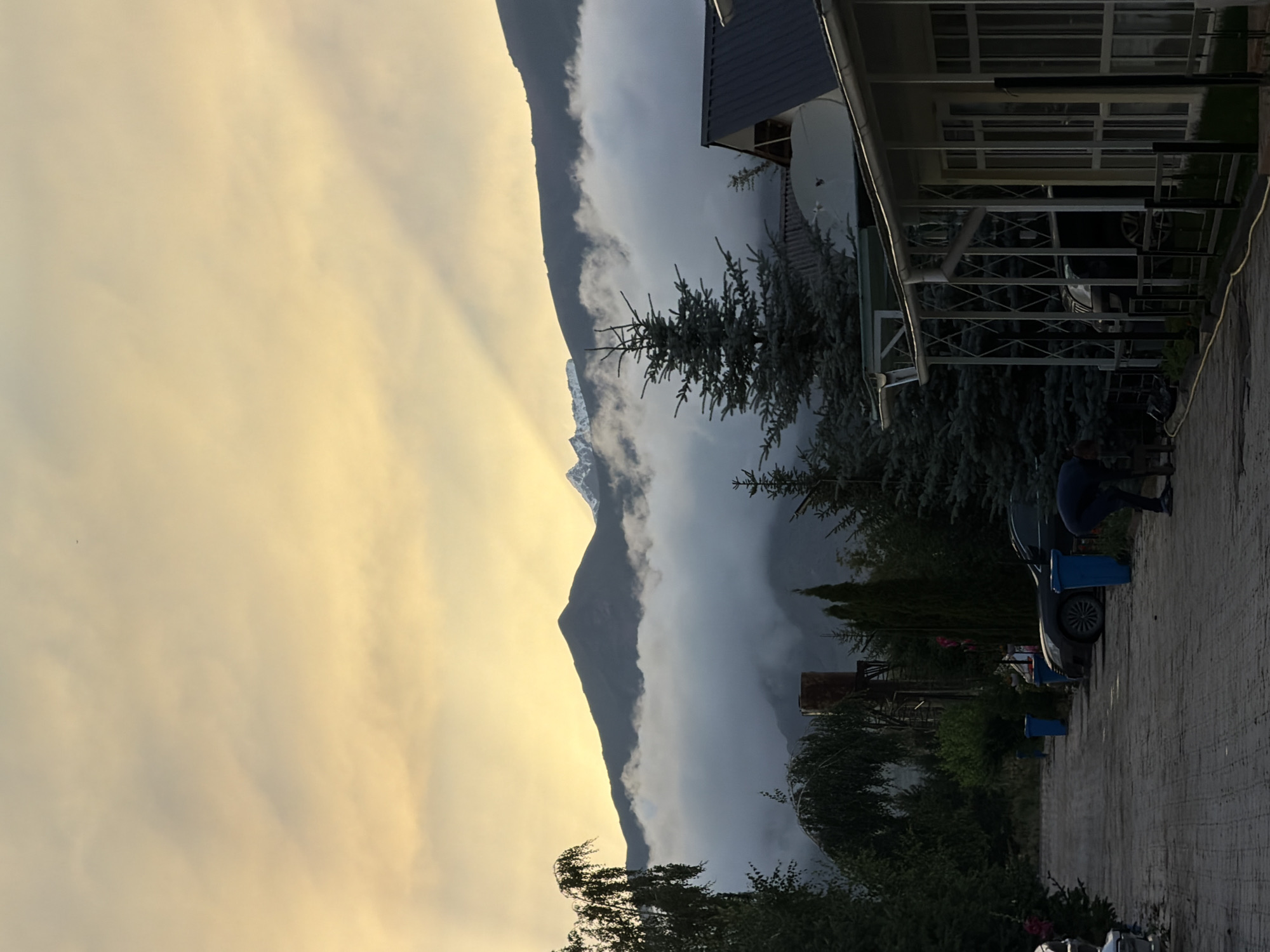 The image depicts a serene morning scene in Çolpon-Ata, Kyrgyzstan, with the dramatic backdrop of mountains partially obscured by low-lying clouds. The sky is illuminated with the soft, warm hues of sunrise, casting ethereal rays of light through the clouds and onto the mountain peaks. In the foreground, a paved pathway leads into the frame, flanked by evergreen trees and several parked vehicles, including a dark-colored sedan and an SUV. A person in dark clothing is seen stooping down on the pathway near the vehicles, appearing to be engaged in an activity on the ground. Adjacent to the pathway, a building with a white exterior and a dark roof is visible, featuring a covered porch supported by slender white pillars. The overall atmosphere suggests a peaceful, rural setting in the early hours of the day.