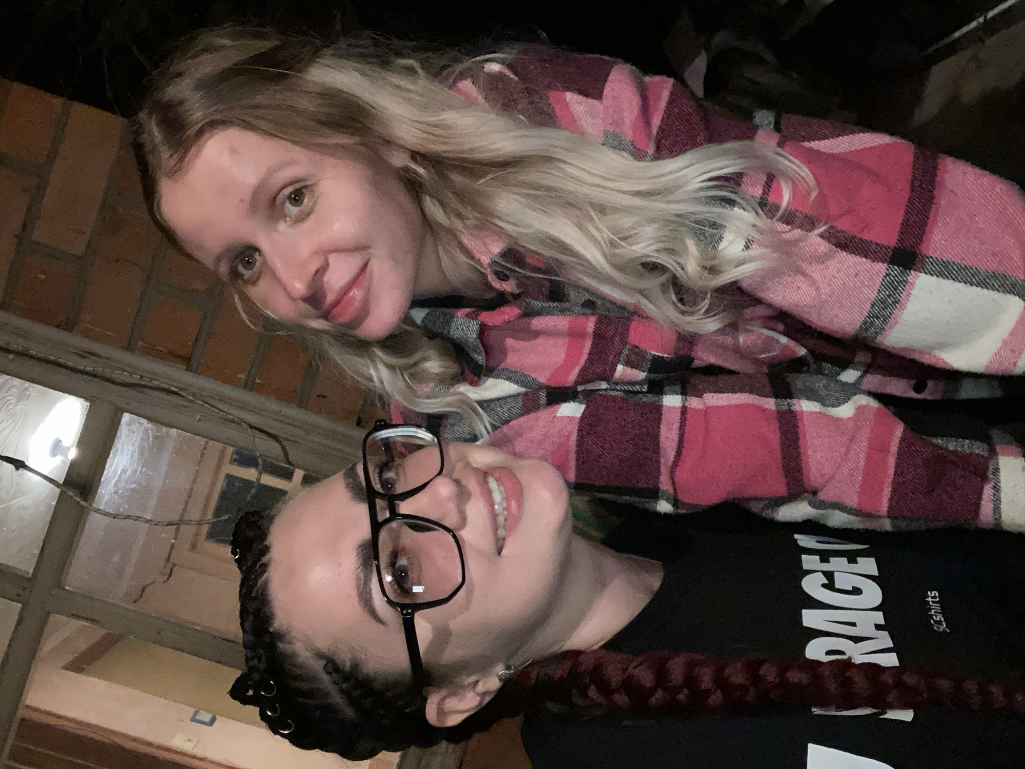 Two women are captured in a close-up, selfie-style photograph. The woman on the left has dark hair styled in braids adorned with small silver rings, wears large, dark-rimmed glasses, and a black t-shirt with white lettering. The woman on the right has long, blonde, wavy hair and is wearing a pink and black plaid shirt. Both women are smiling and appear to be outdoors, with a brick wall and a wooden window frame visible in the background. The lighting suggests it is dusk or nighttime. The location is identified as Солнечная Поляна, Russia.