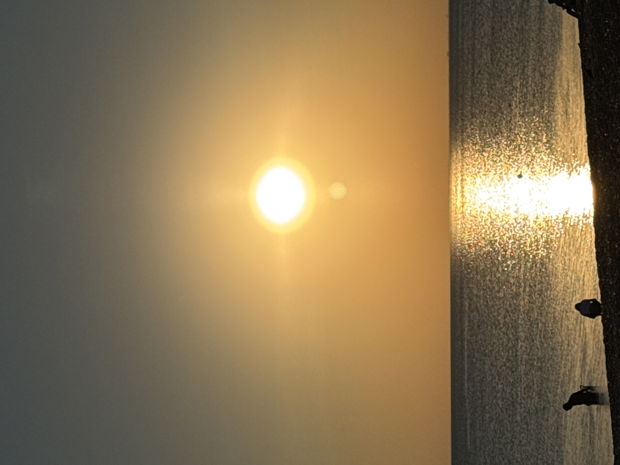 A vertical, outdoor, close-up shot captures the setting sun over a calm body of water at the Novofedorovka beach in Ukraine. The sun, a bright white orb with visible rays, is positioned in the upper-middle of the frame, casting a golden hue across the sky and reflecting intensely on the water's surface. Two silhouettes of people are visible on the pebbled shore in the foreground. One, a solitary figure, stands partially submerged in the shallow water on the left side, facing away from the camera. To the right of this figure, a second silhouette, appearing smaller and possibly seated or crouched, is also present on the shore. The water is a dark gray, contrasting with the bright reflection of the sun. The horizon line is clearly defined, separating the water from the sky. The overall atmosphere is serene and tranquil.