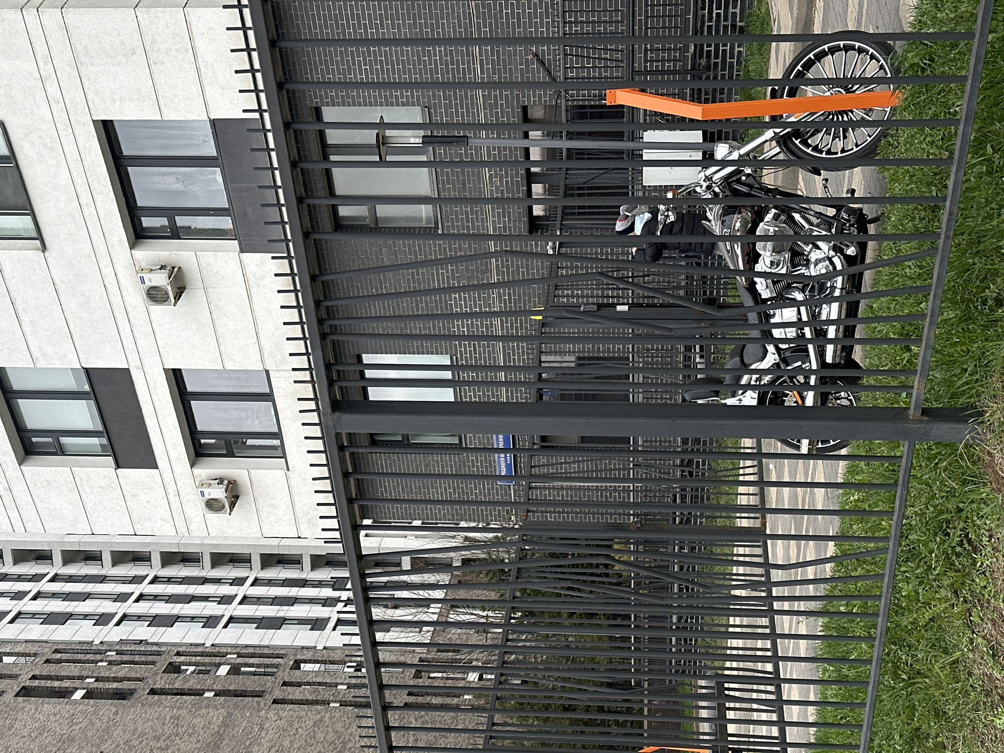 The image shows a black metal fence in the foreground with a view of buildings and a motorcycle behind it. The motorcycle is chrome-colored with an orange strap attached. The buildings in the background have a mix of white and gray brick facades with windows and air conditioning units. A street sign is visible behind the fence indicating that the location is in Moscow, Russia, specifically at "Klyazmkoo Street." Green grass is visible on the right side of the image.