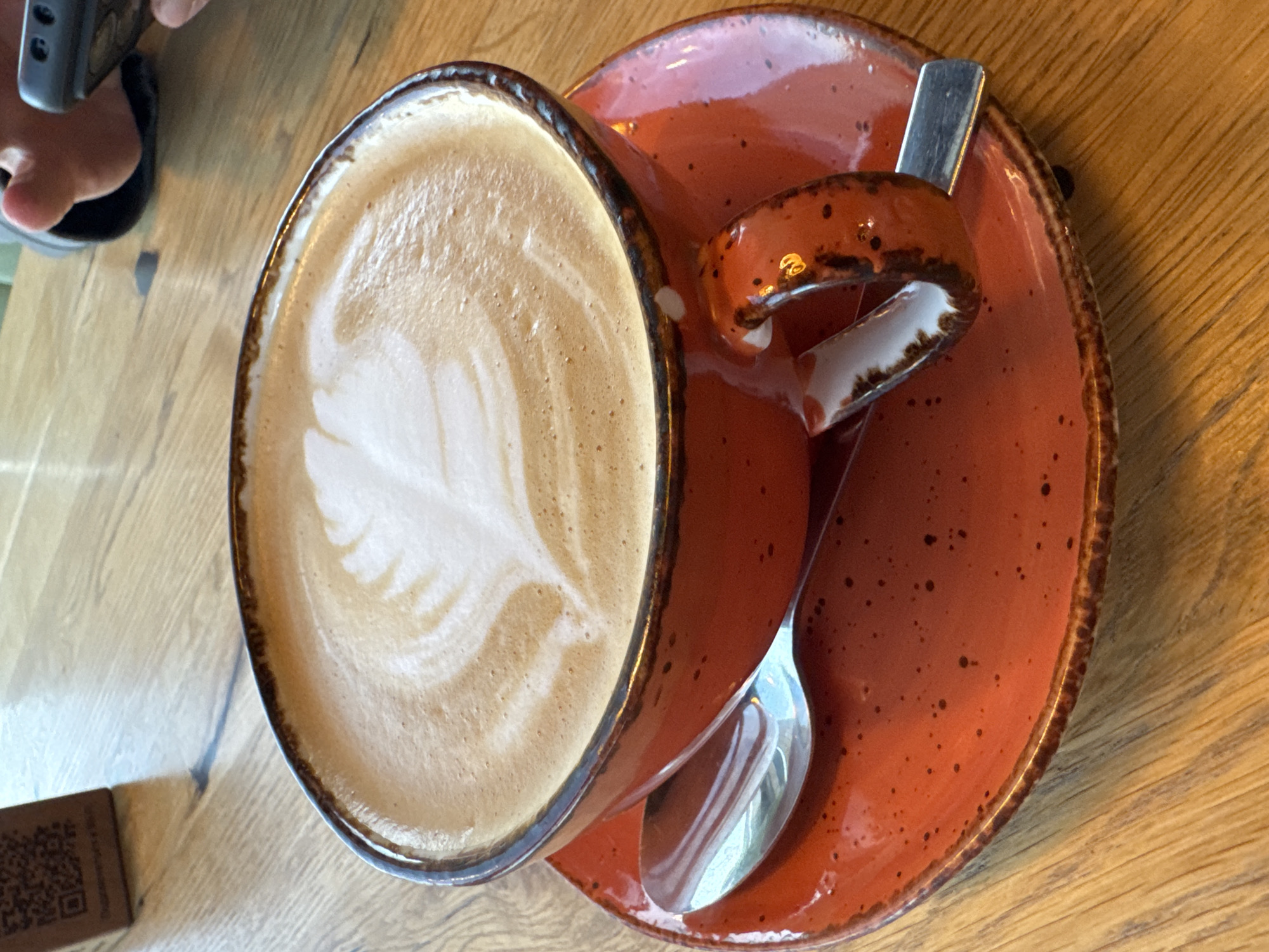 A close-up, eye-level shot shows a vibrant orange ceramic cup and saucer filled with latte art coffee, set on a wooden table. The latte art is a white, leaf-like design atop the frothy, light brown coffee. A silver spoon rests on the saucer. The cup has a textured, dark brown rim and a uniquely shaped handle with a glossy, speckled finish. In the background, a QR code sign is visible on the left, and to the right, a person's hands are partially visible, holding a mobile phone. The lighting suggests an indoor setting. This scene is located in the city of Kurkijoki, Russia.
