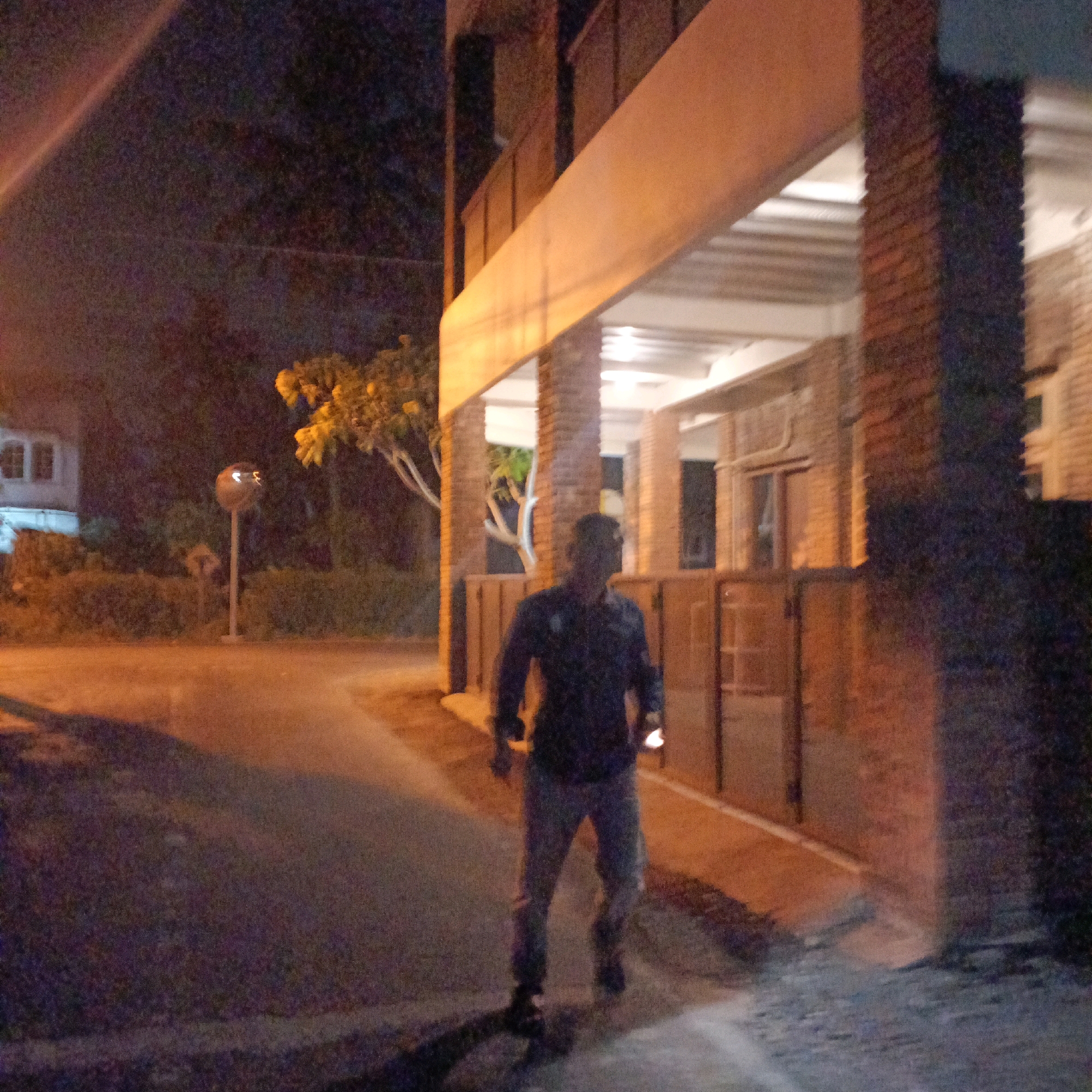 In Salatiga, Indonesia, a man is walking on a street at night. He is wearing a dark, long-sleeved shirt and light-colored pants. The street is paved and appears to be dimly lit by artificial lights from nearby buildings. To the right of the man is a building with a brick facade and a covered walkway supported by brick columns. Yellowish light emanates from the covered area. To the left of the man, a tree with yellow flowers is visible, and further in the background, a house with two windows can be seen. A street lamp with a reflective surface is also present on the left side of the image. The overall scene is dark, with the primary light sources being artificial illumination.