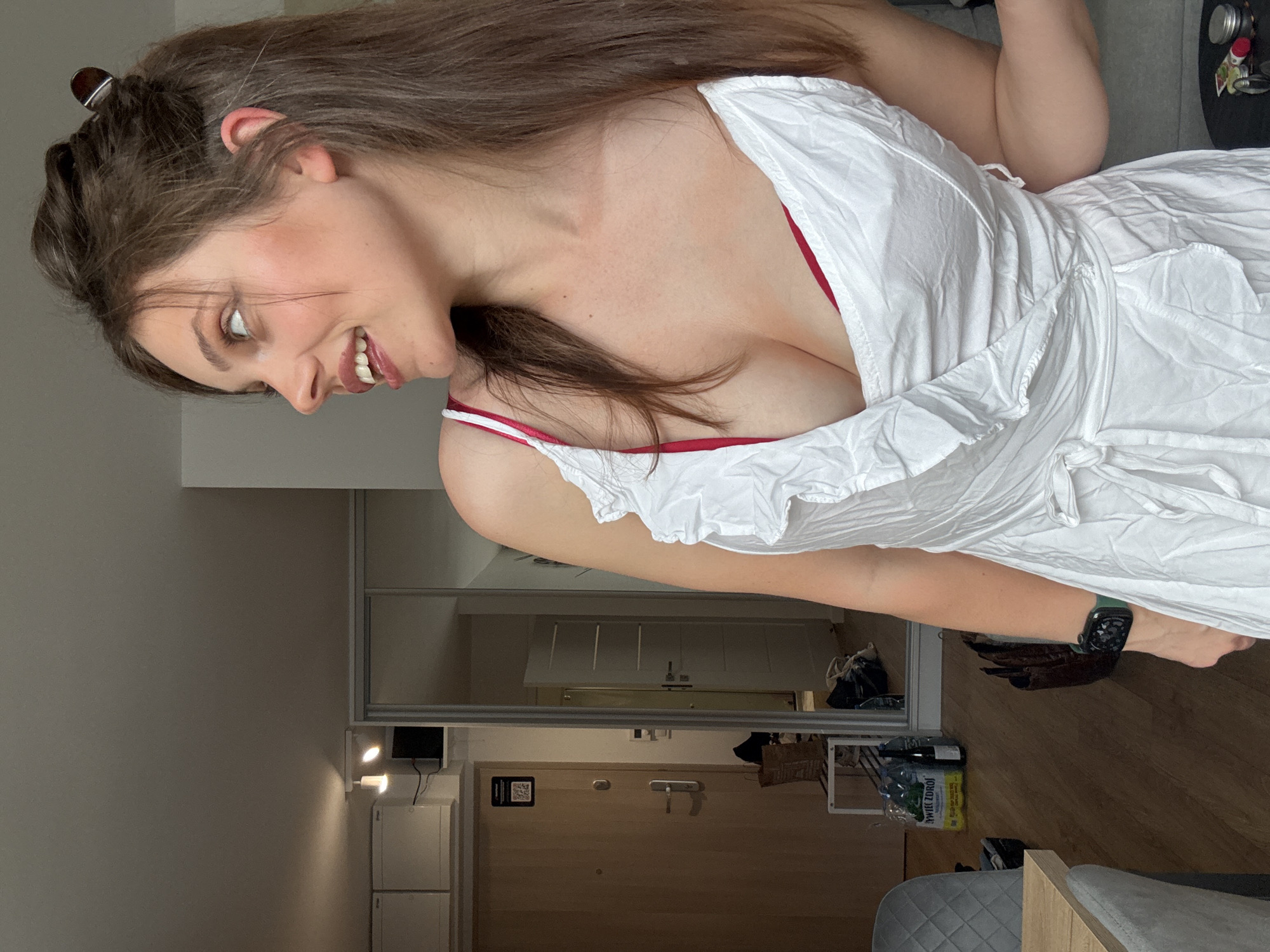 The image shows a woman in an apartment. She is wearing a white dress and a red bra. Her hair is brown and pulled up in a bun. She is looking to the side and smiling. In the background, there is a door, a mirror, and some furniture. The floor is wooden. There are bottles of water on the floor. The scene appears to be in an urban apartment, not in Szklarska Poręba, Poland, as there are no visible landmarks or context to suggest that specific location.